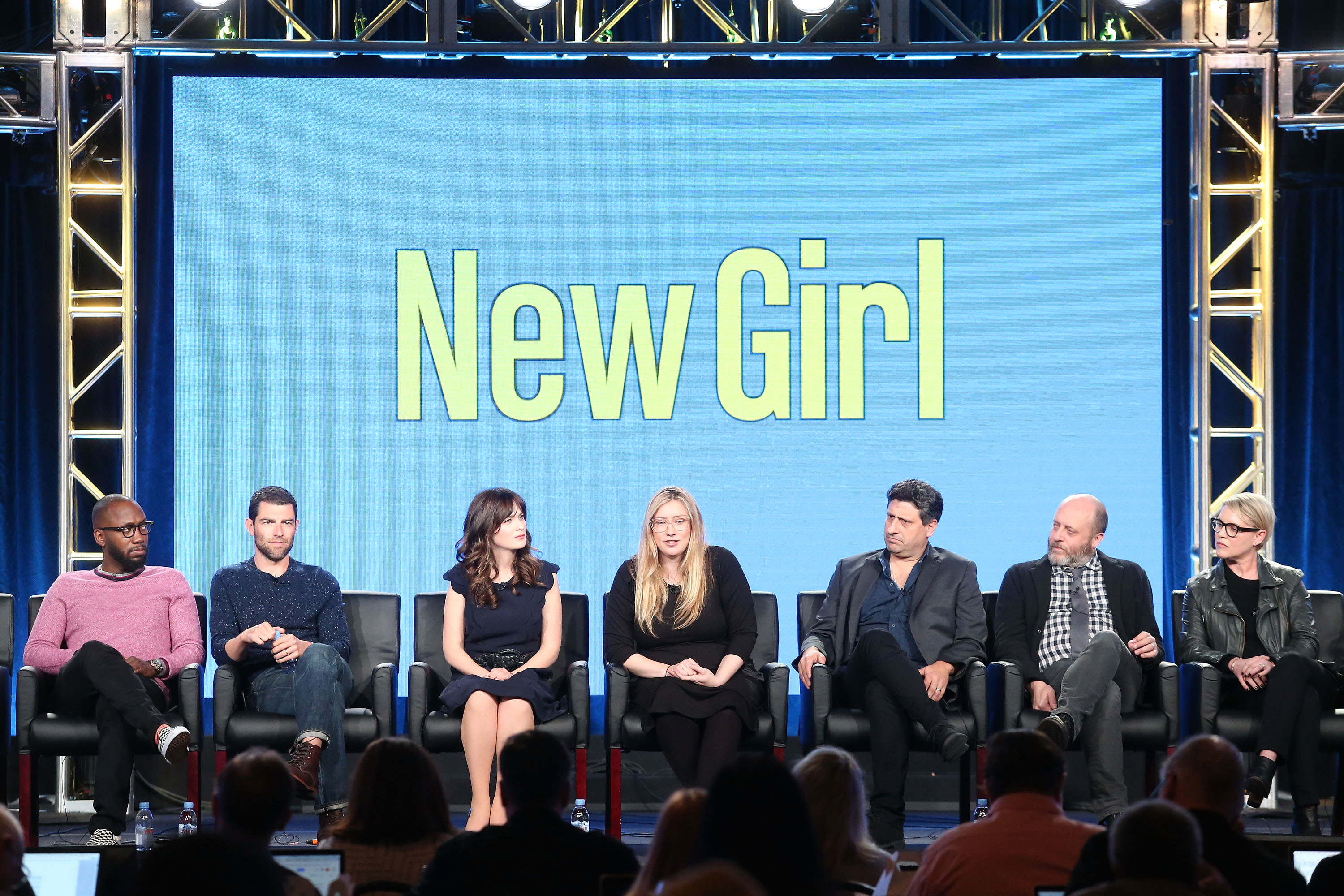 Click to open the image gallery Seven cast and crew members of New Girl seated on a panel stage in front of a blue screen reading 'New Girl' at the 2018 Winter TCA Press Tour.