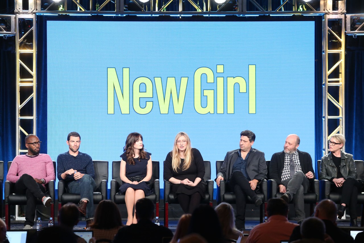 Seven cast and crew members of New Girl seated on a panel stage in front of a blue screen reading 'New Girl' at the 2018 Winter TCA Press Tour.