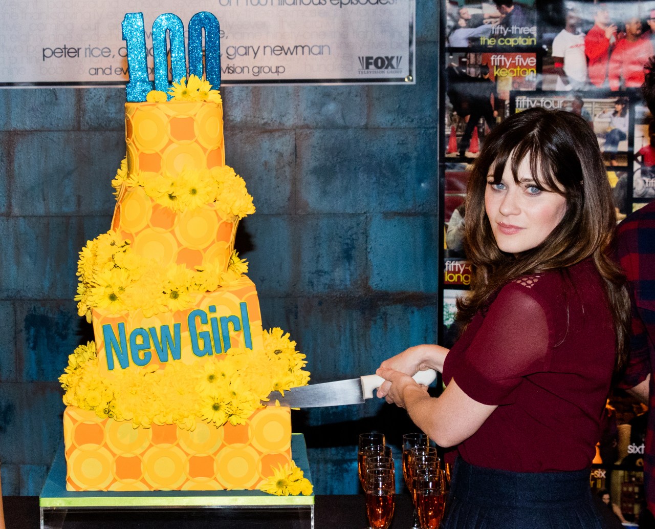 A woman in a burgundy top slices a large yellow tiered cake decorated with yellow flowers and a blue '100' topper reading 'New Girl.'