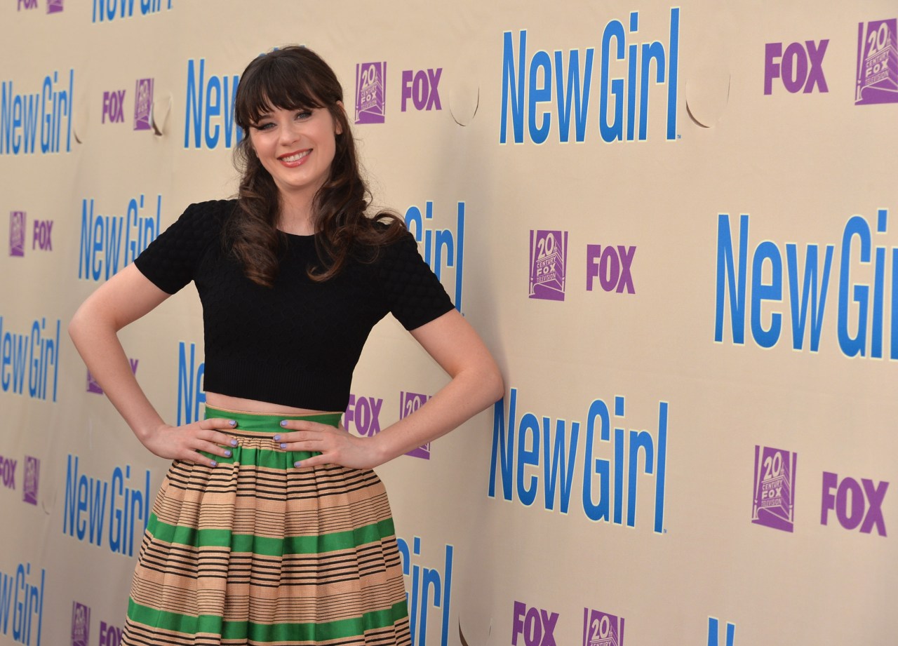A smiling woman with dark hair and bangs stands in front of a New Girl and FOX step-and-repeat backdrop, wearing a black crop top and striped skirt.