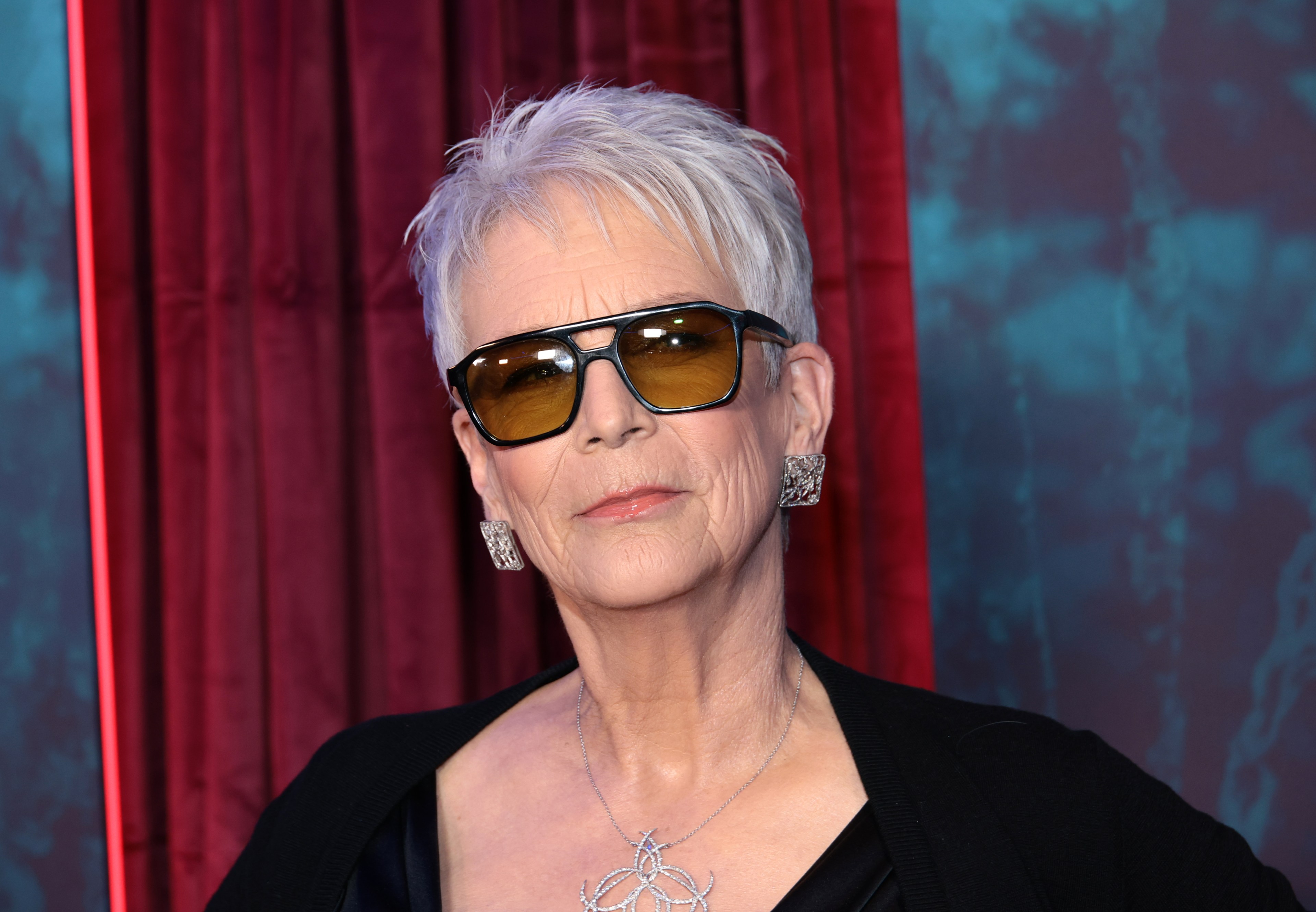 Jamie Lee Curtis wearing black sunglasses, a black top, silver earrings, and a silver necklace at a red-carpet premiere event.