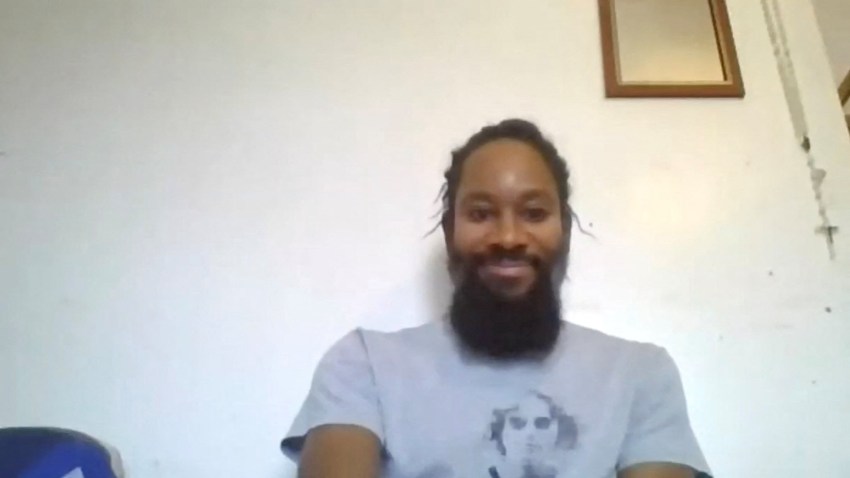 A bearded man with braided hair smiles while seated indoors in front of a white wall, wearing a gray graphic t-shirt.