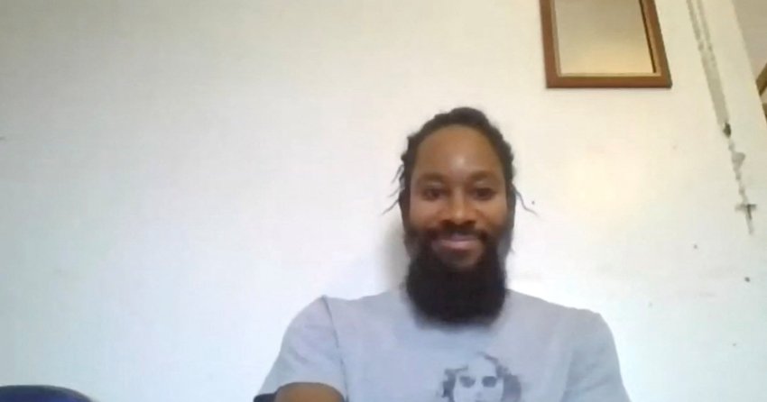 Man with braided hair and a full beard wearing a gray graphic t-shirt, seated in front of a white wall with a framed picture in the background.