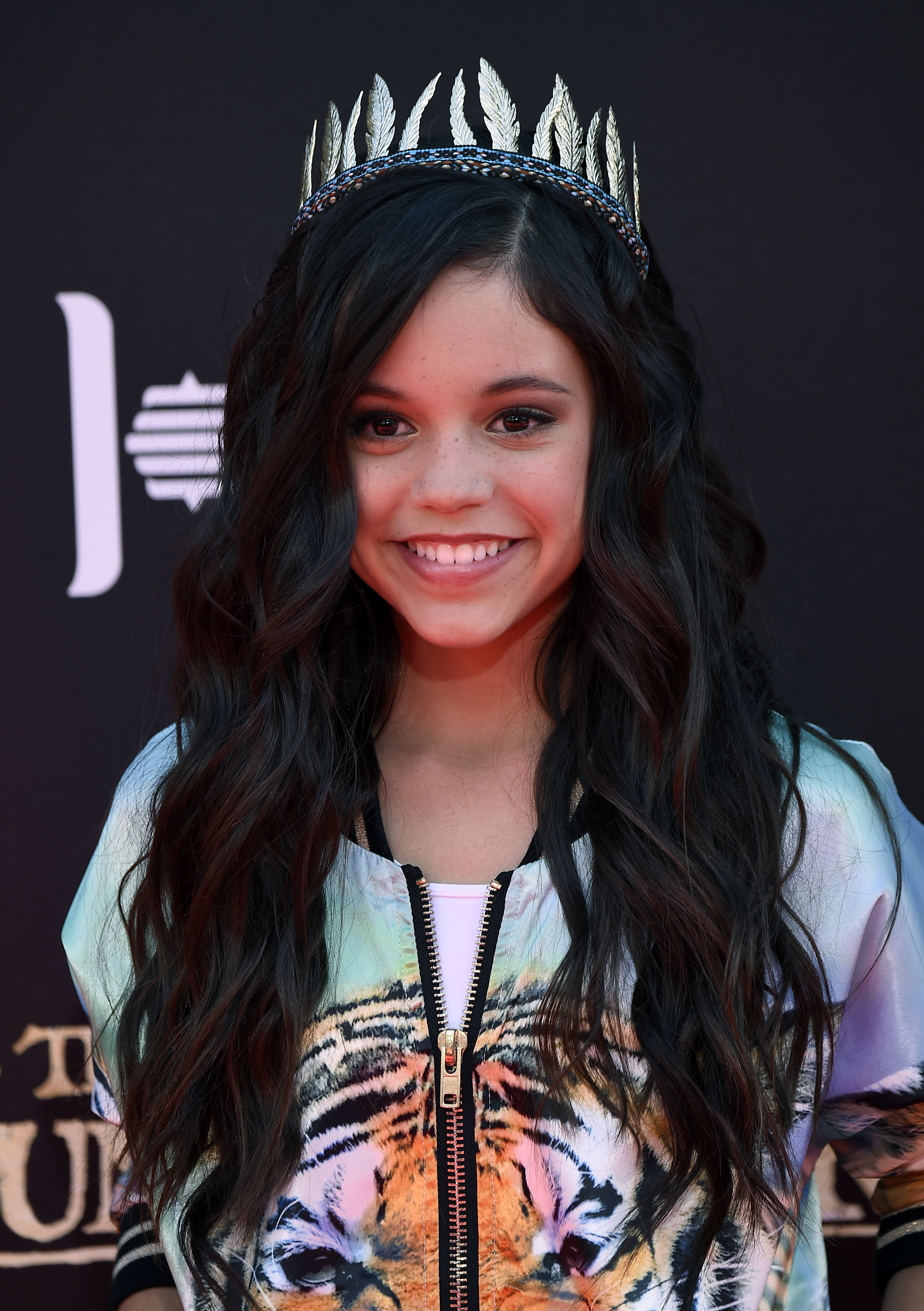 Young Jenna Ortega smiling at The Jungle Book Disney premiere, wearing a silver feather crown headband and a tiger-print zip-up jacket.