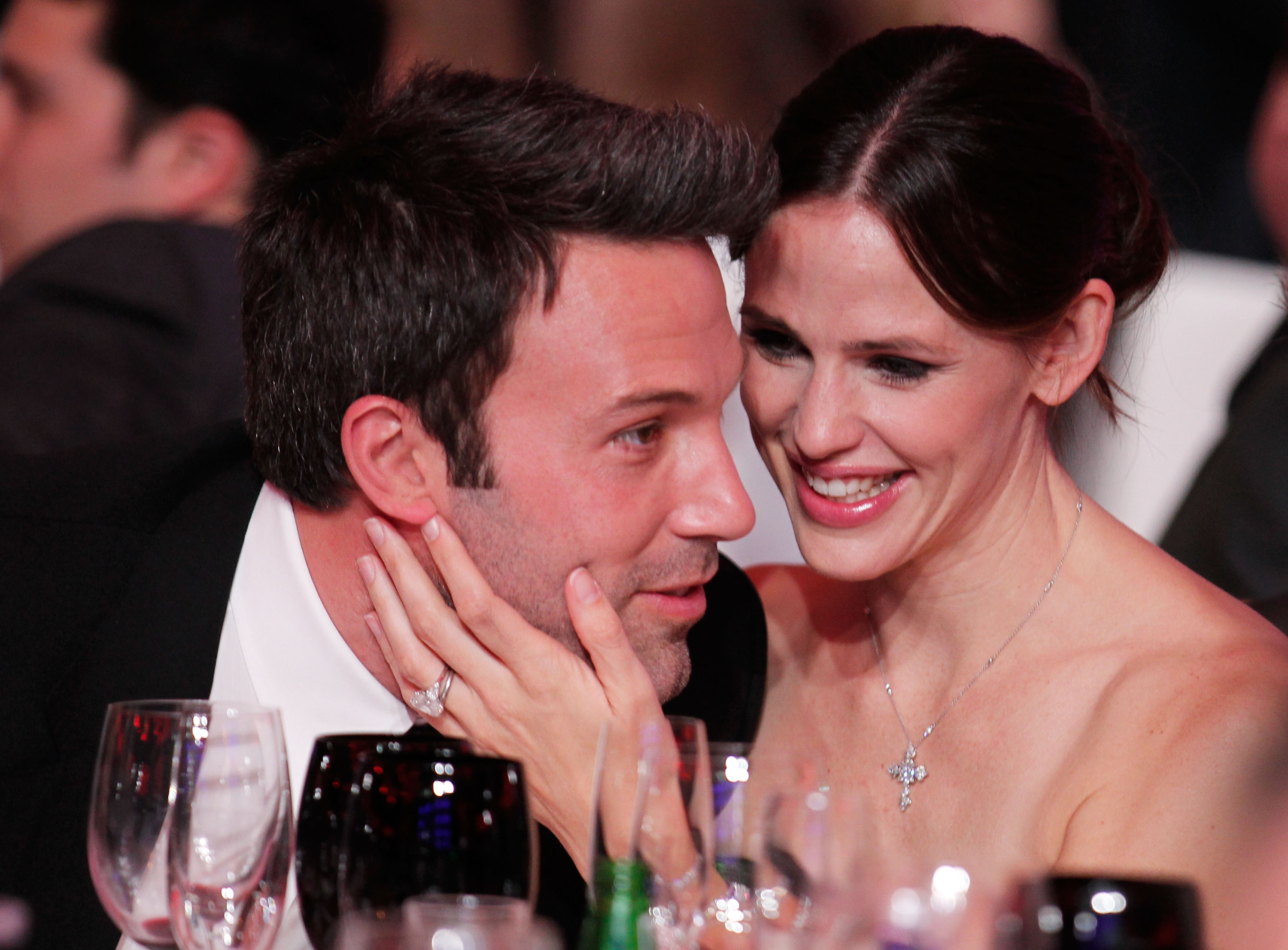 Ben Affleck and Jennifer Garner lean in close to each other at a gala dinner table, smiling, with wine glasses in the foreground.