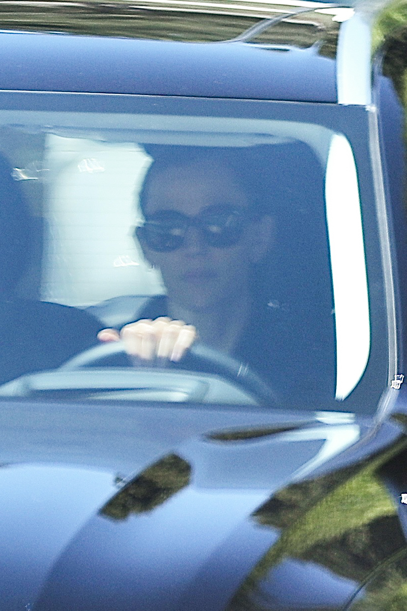 A person wearing sunglasses seen through the windshield of a blue SUV, hand on the steering wheel.