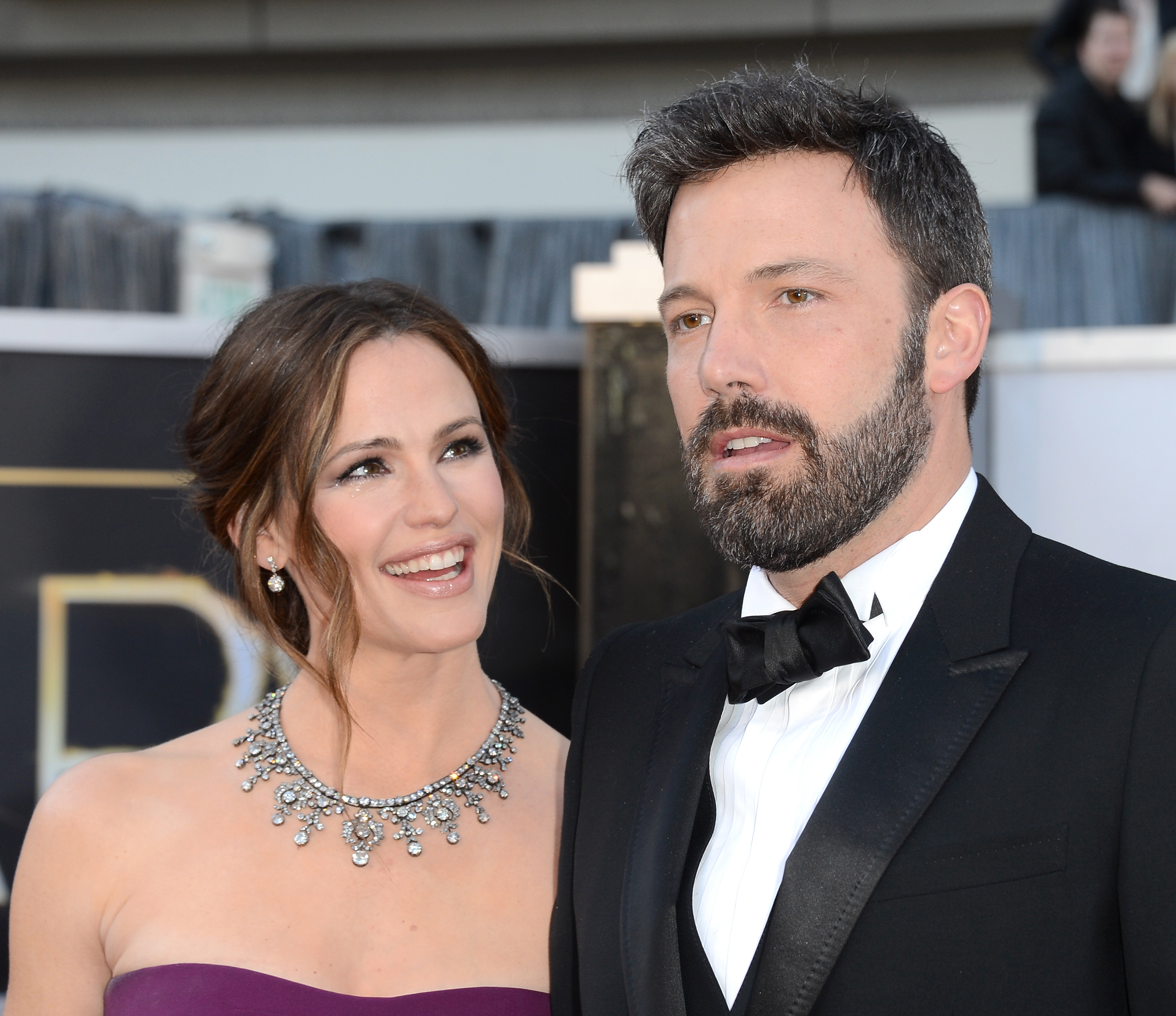 Jennifer Garner in a purple strapless gown with a diamond necklace smiles beside Ben Affleck in a black tuxedo on a red carpet.