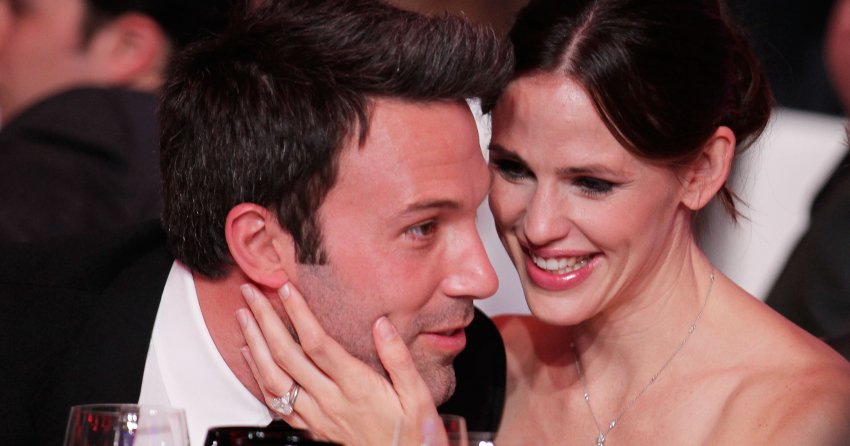 A man and woman lean close together at a formal dinner table, the woman smiling and touching the man's face affectionately, wine glasses in the foreground.