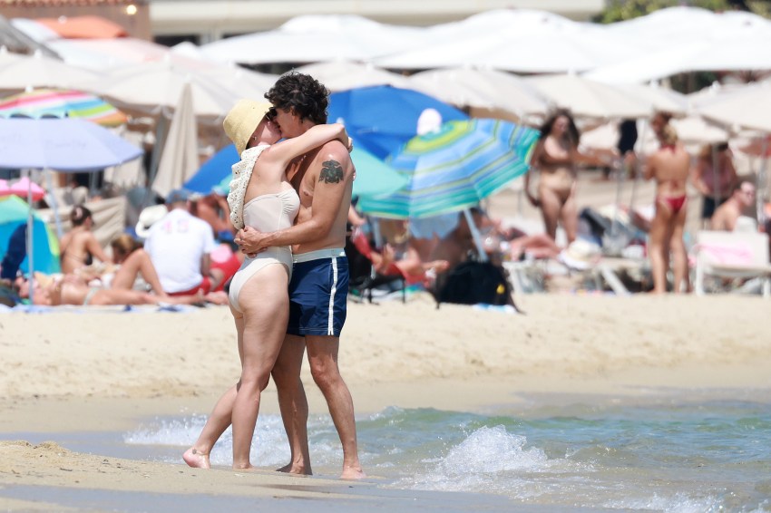 A couple embracing and kissing on a crowded sandy beach, surrounded by umbrellas and sunbathers, the man shirtless in blue swim trunks, the woman in a nude swimsuit and yellow sun hat.