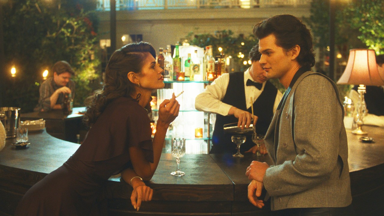 A woman holding a cigarette leans on a dimly lit bar talking to a young man in a gray jacket, with a bartender in a bow tie behind them.