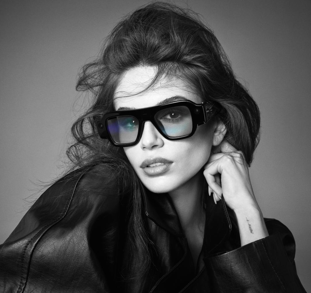 Black-and-white portrait of a dark-haired woman wearing oversized black smart glasses and a leather jacket, hand raised to her chin.