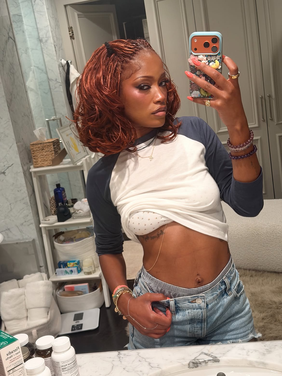 Woman with auburn braids taking a bathroom mirror selfie in a tied-up white and navy baseball tee and low-rise jeans.