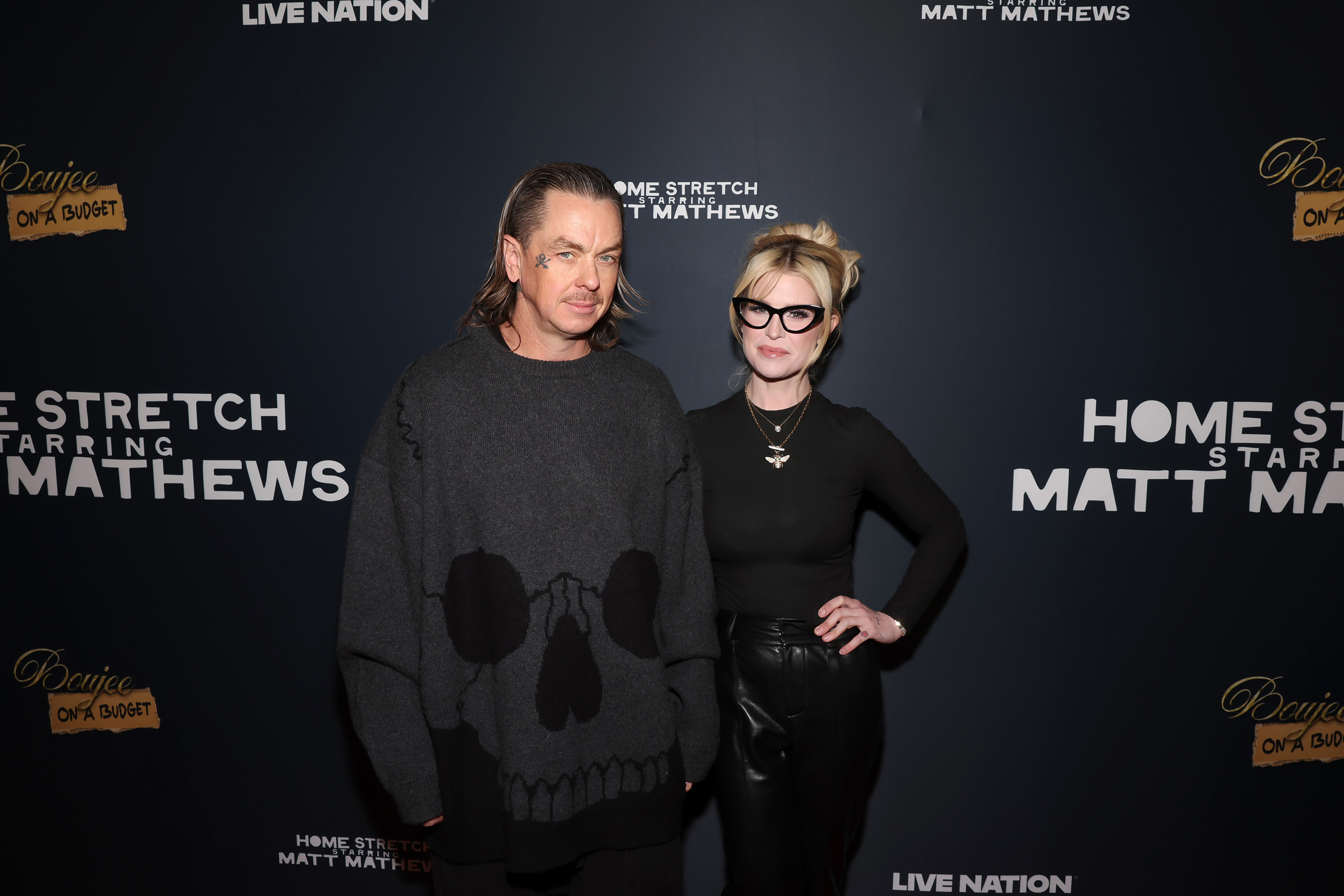 Sid Wilson in a skull-print sweater and Kelly Osbourne in all black with cat-eye glasses pose at a Live Nation event backdrop.