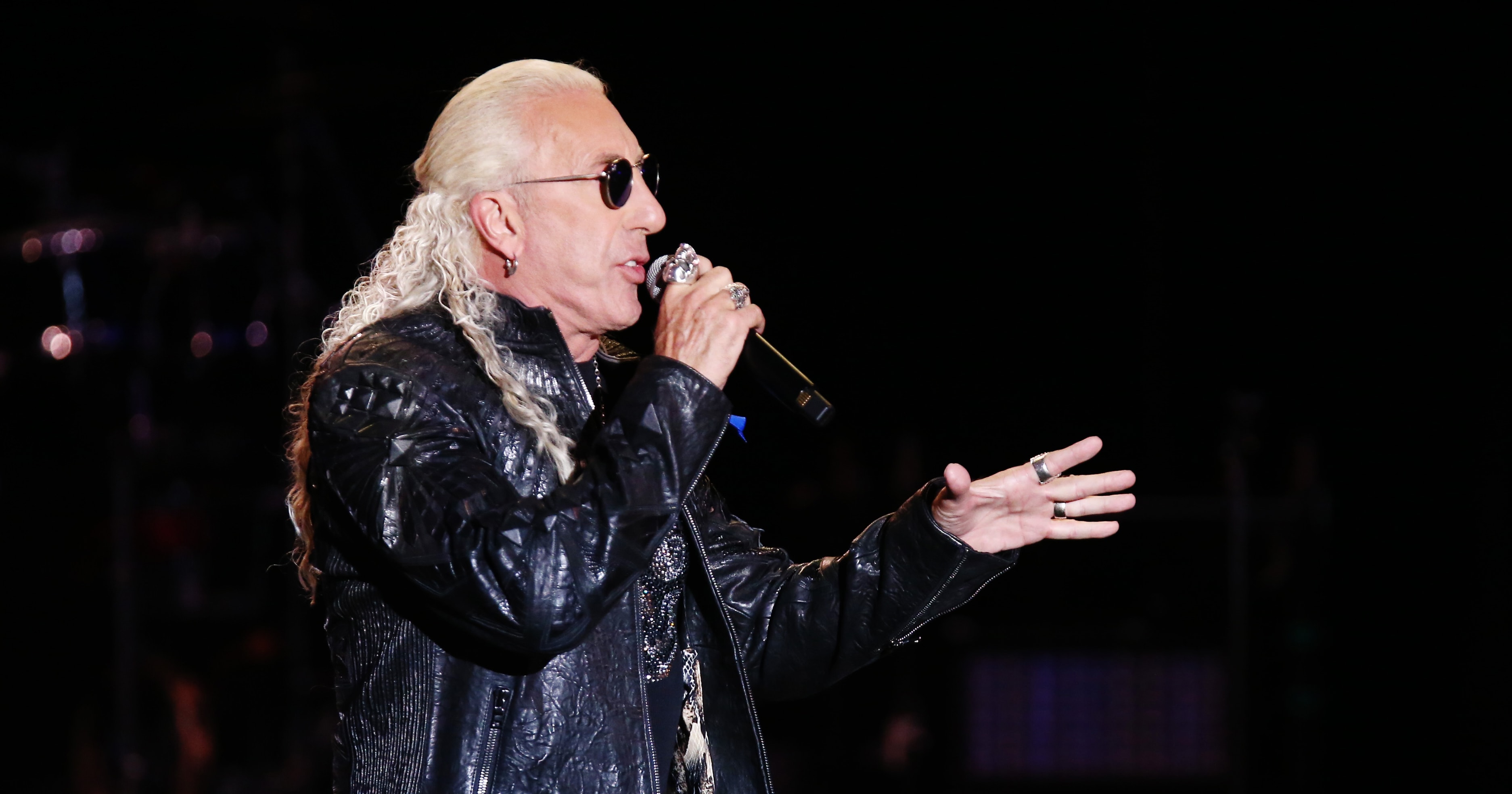 Dee Snider singing into a microphone onstage, wearing a black leather jacket and sunglasses with long blonde curly hair.