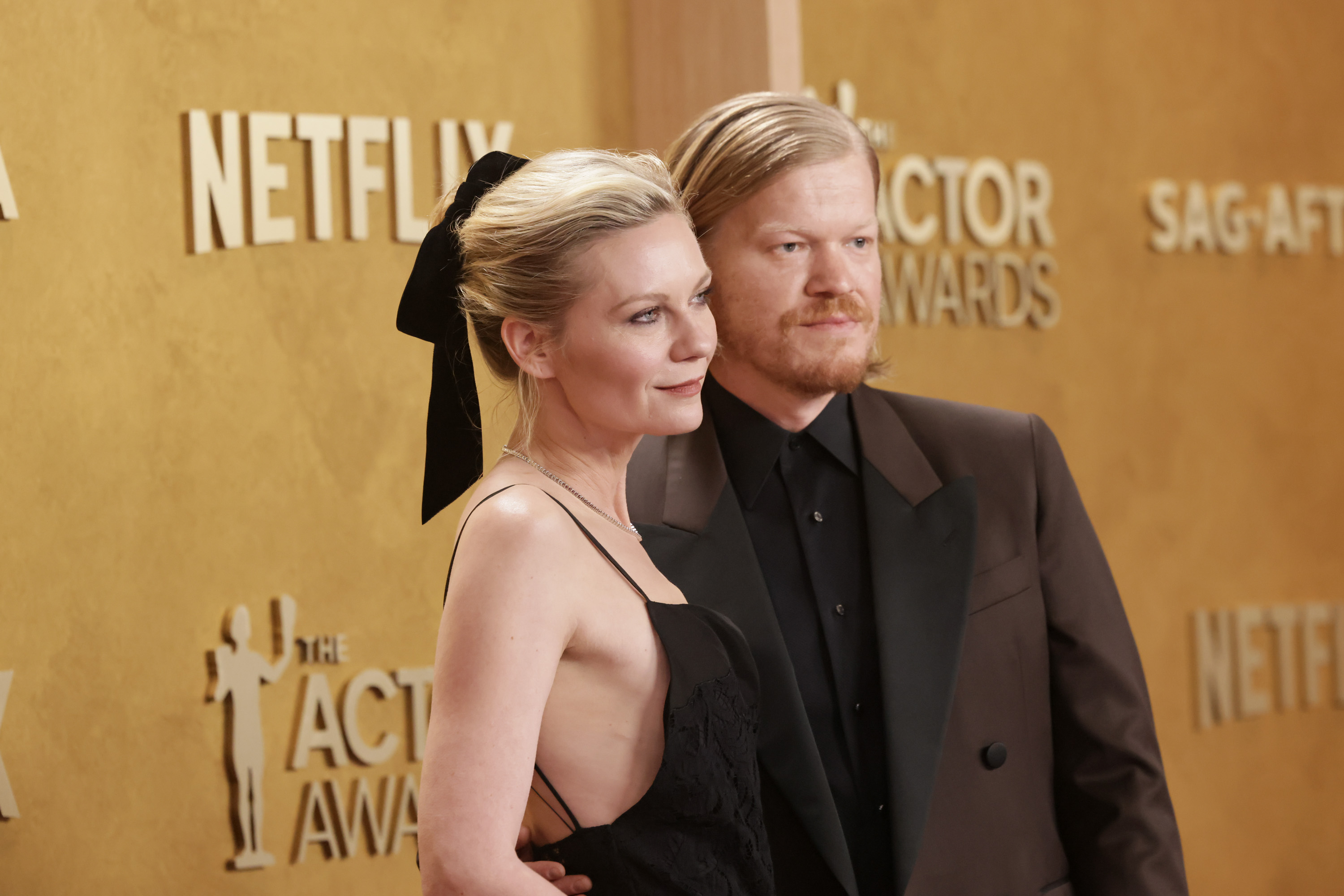 Kirsten Dunst in a black gown with a black bow in her hair stands beside Jesse Plemons in a brown suit at the SAG Awards red carpet.