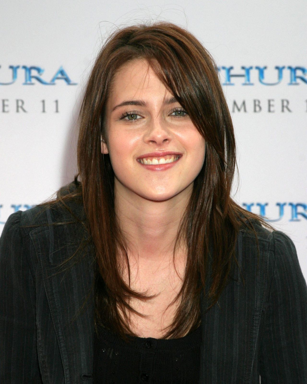 Kristen Stewart smiling with long brown hair at a movie premiere, wearing a dark blazer against a branded step-and-repeat backdrop.
