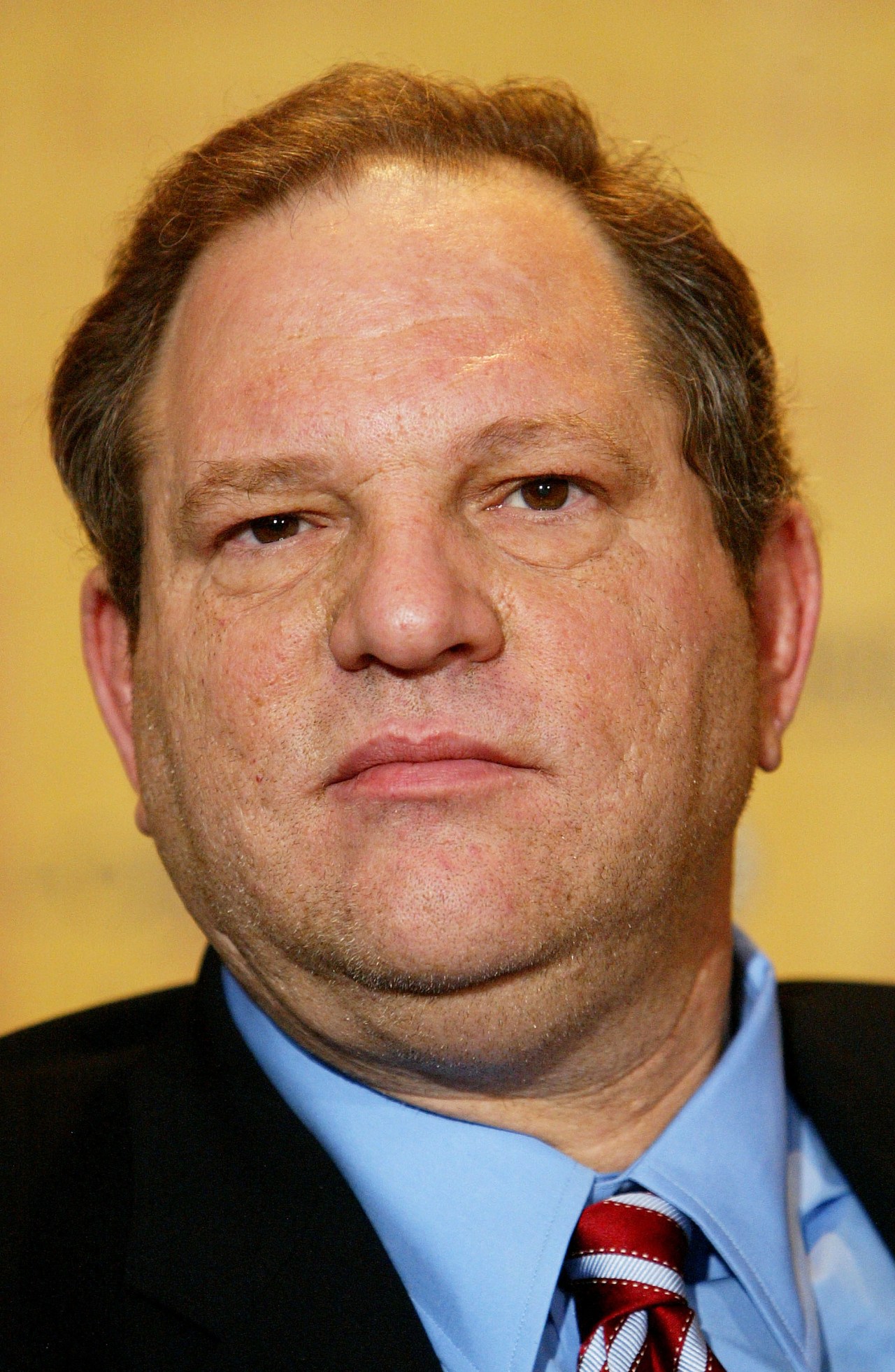 Close-up of a heavyset middle-aged man in a black suit, blue shirt, and red striped tie against a yellow background.