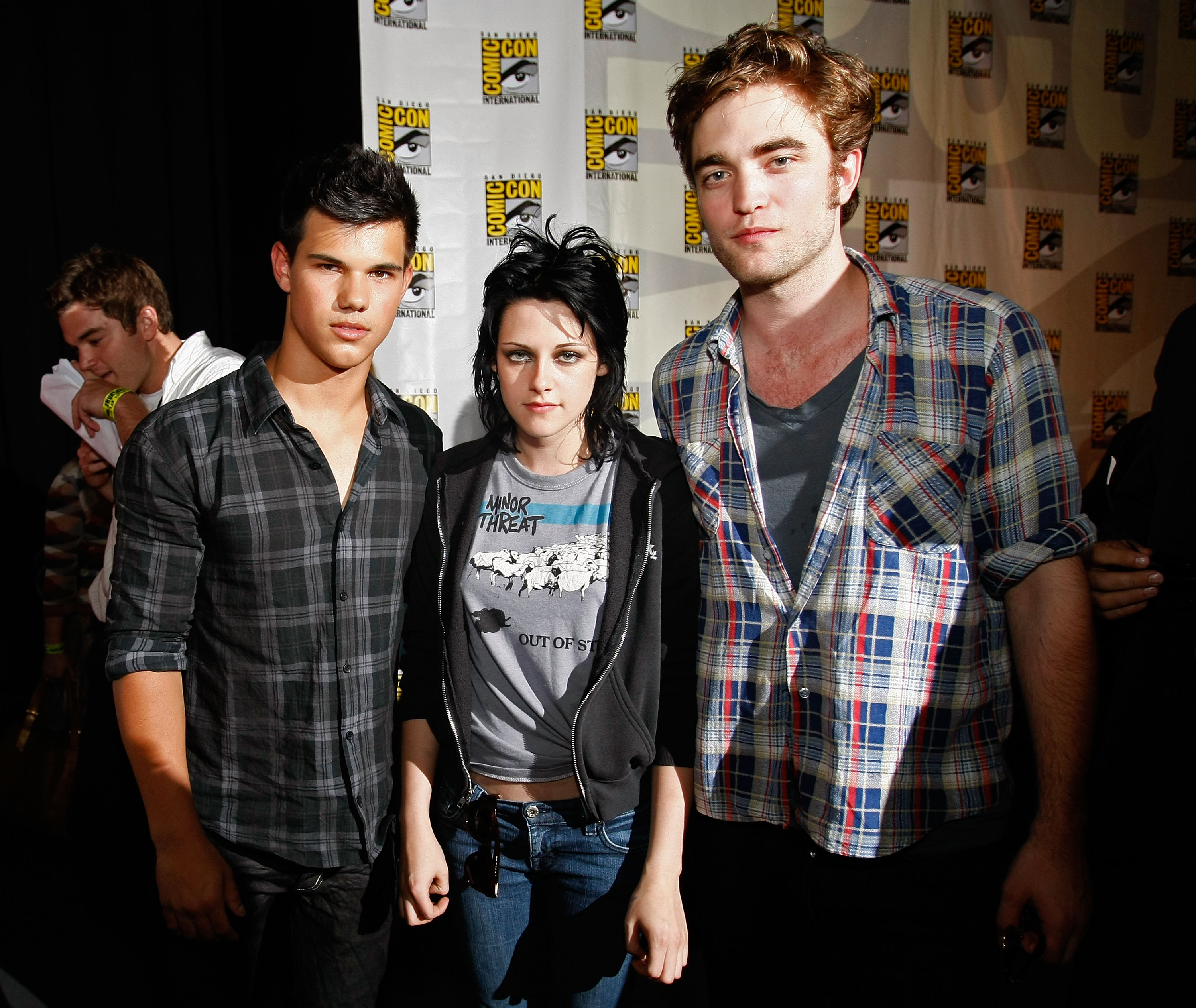 Taylor Lautner, Kristen Stewart, and Robert Pattinson standing together in front of a San Diego Comic-Con step-and-repeat backdrop in 2009.