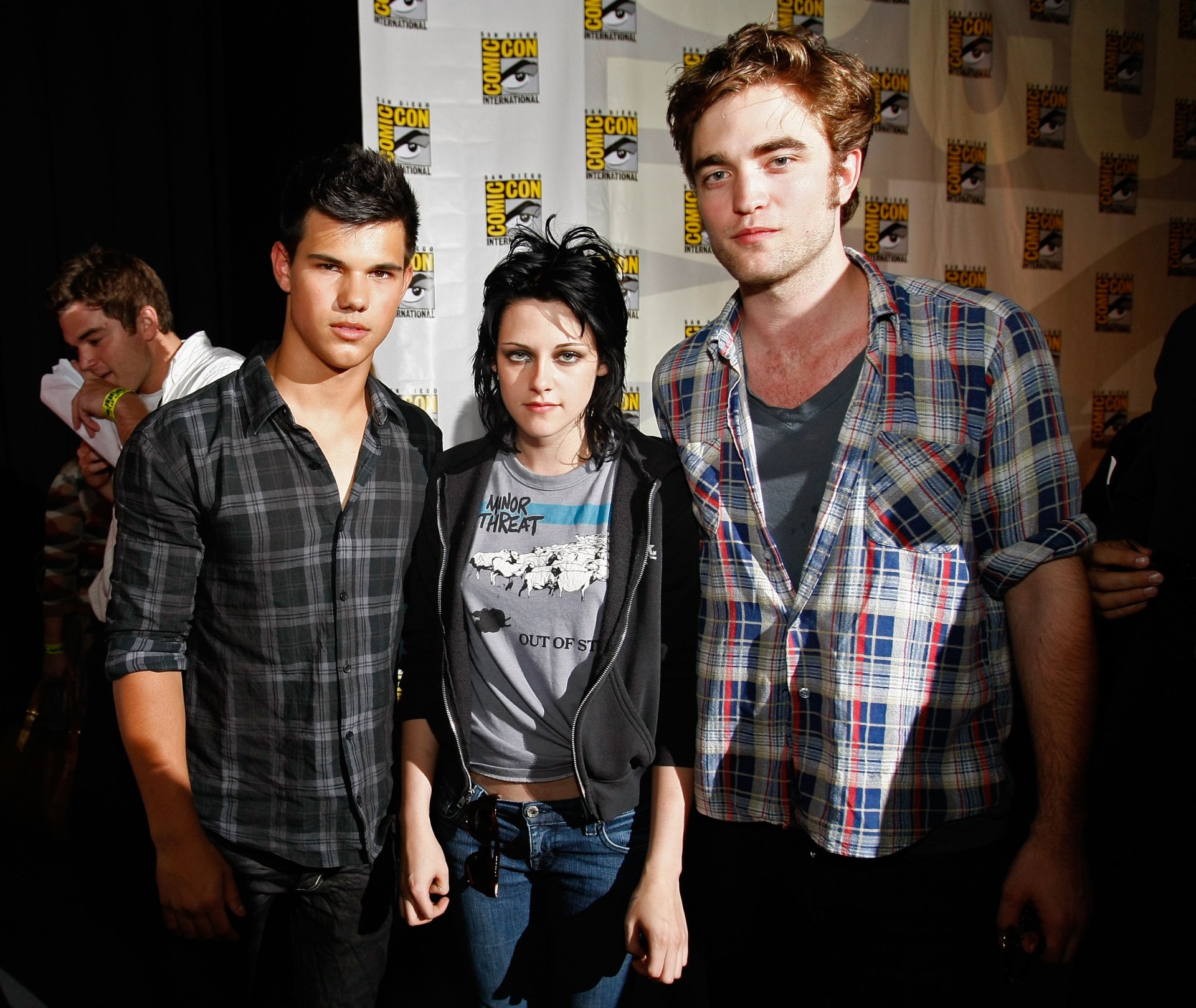 Taylor Lautner, Kristen Stewart, and Robert Pattinson standing together in front of a San Diego Comic-Con step-and-repeat backdrop in 2009.