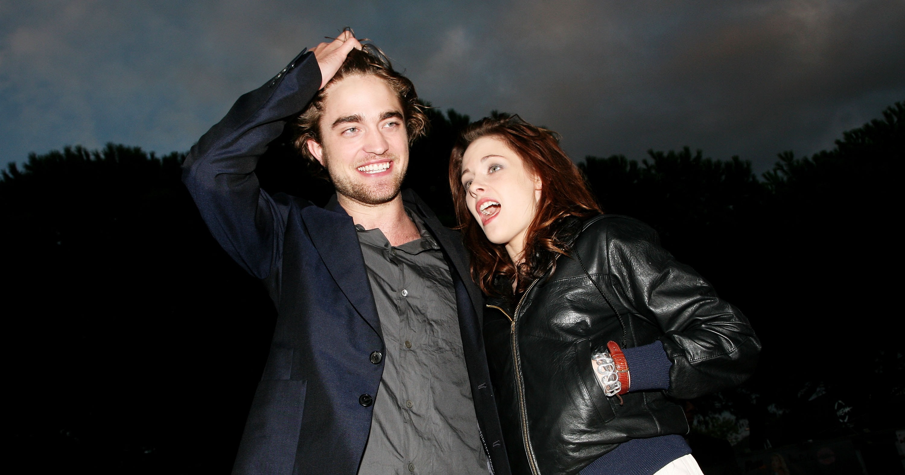 A young man in a blazer laughs while touching his hair as a red-haired woman in a leather jacket leans in beside him at an outdoor evening event.