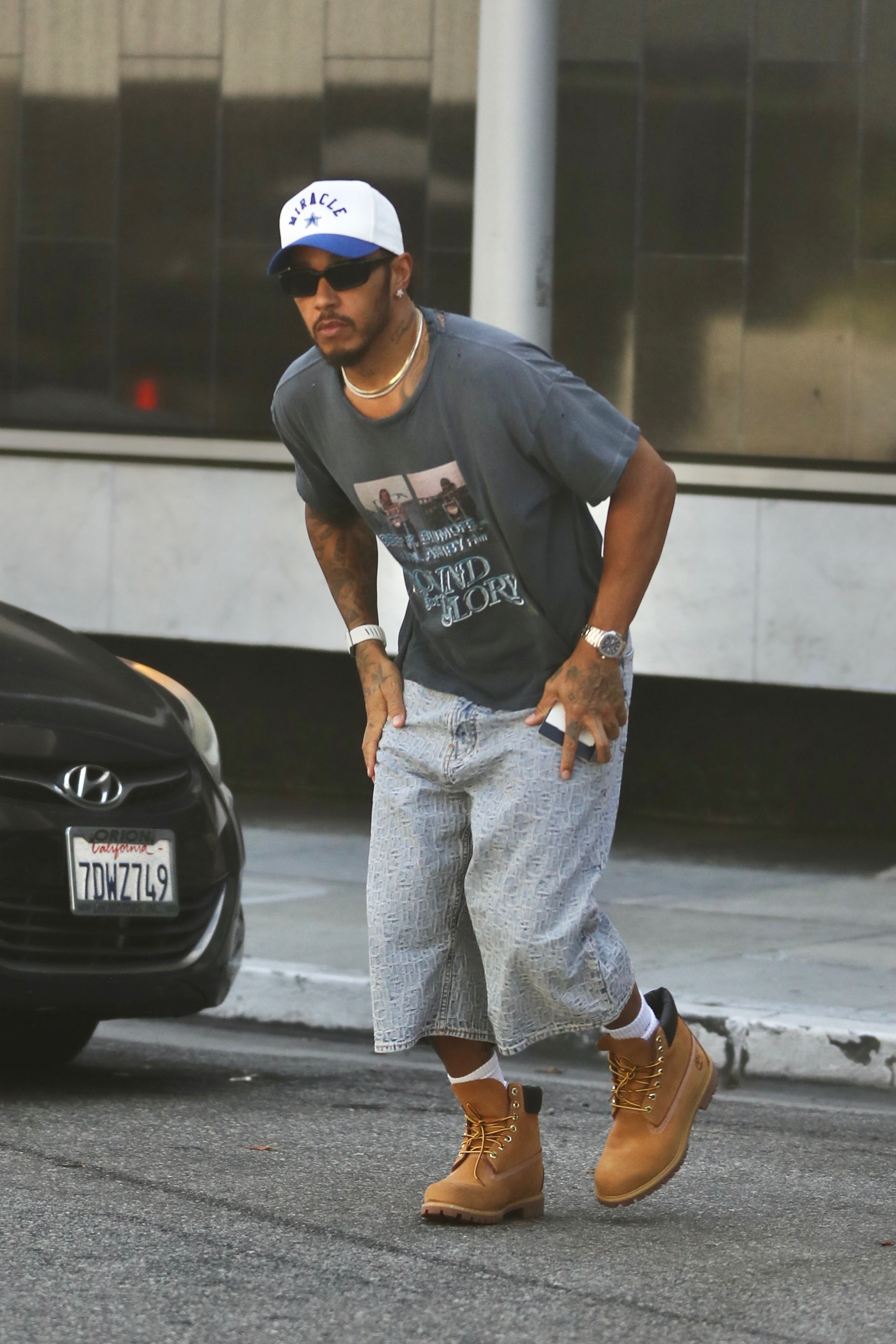 Man in sunglasses, white and blue trucker cap, gray graphic tee, wide-leg denim shorts, and tan Timberland boots bending forward on a sidewalk next to a black car.