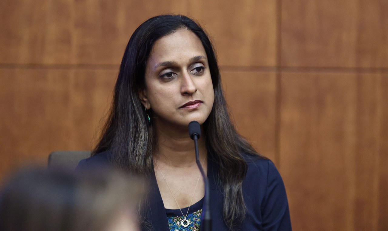 Woman with long dark hair in a navy blazer seated at a witness stand microphone, looking to the side with a neutral expression.