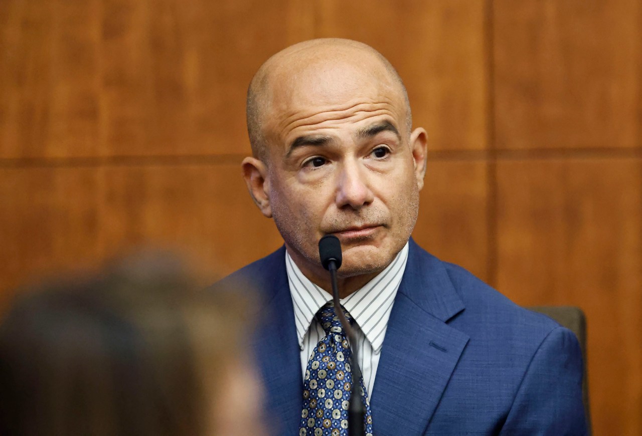 Bald man in a blue suit and patterned tie seated at a witness stand microphone, looking toward the camera with a tense expression.
