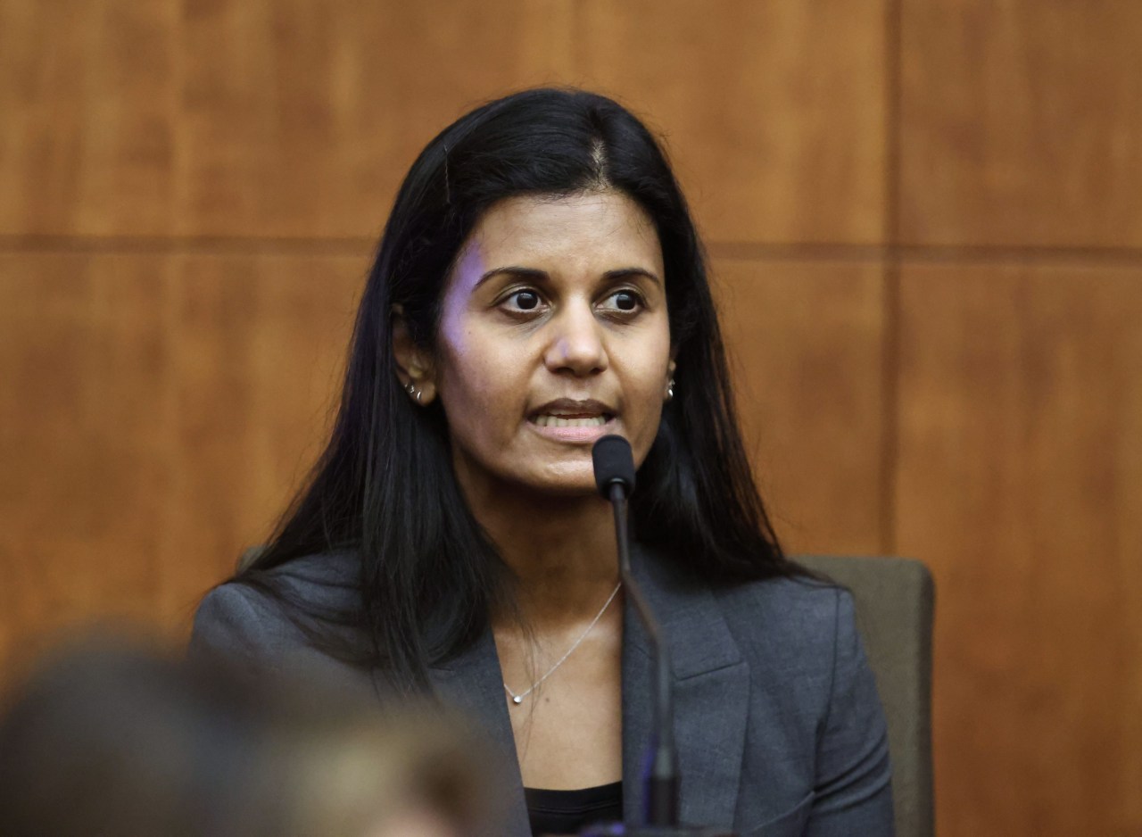Woman with long black hair in a gray blazer speaking into a microphone at a witness stand in a wood-paneled courtroom.