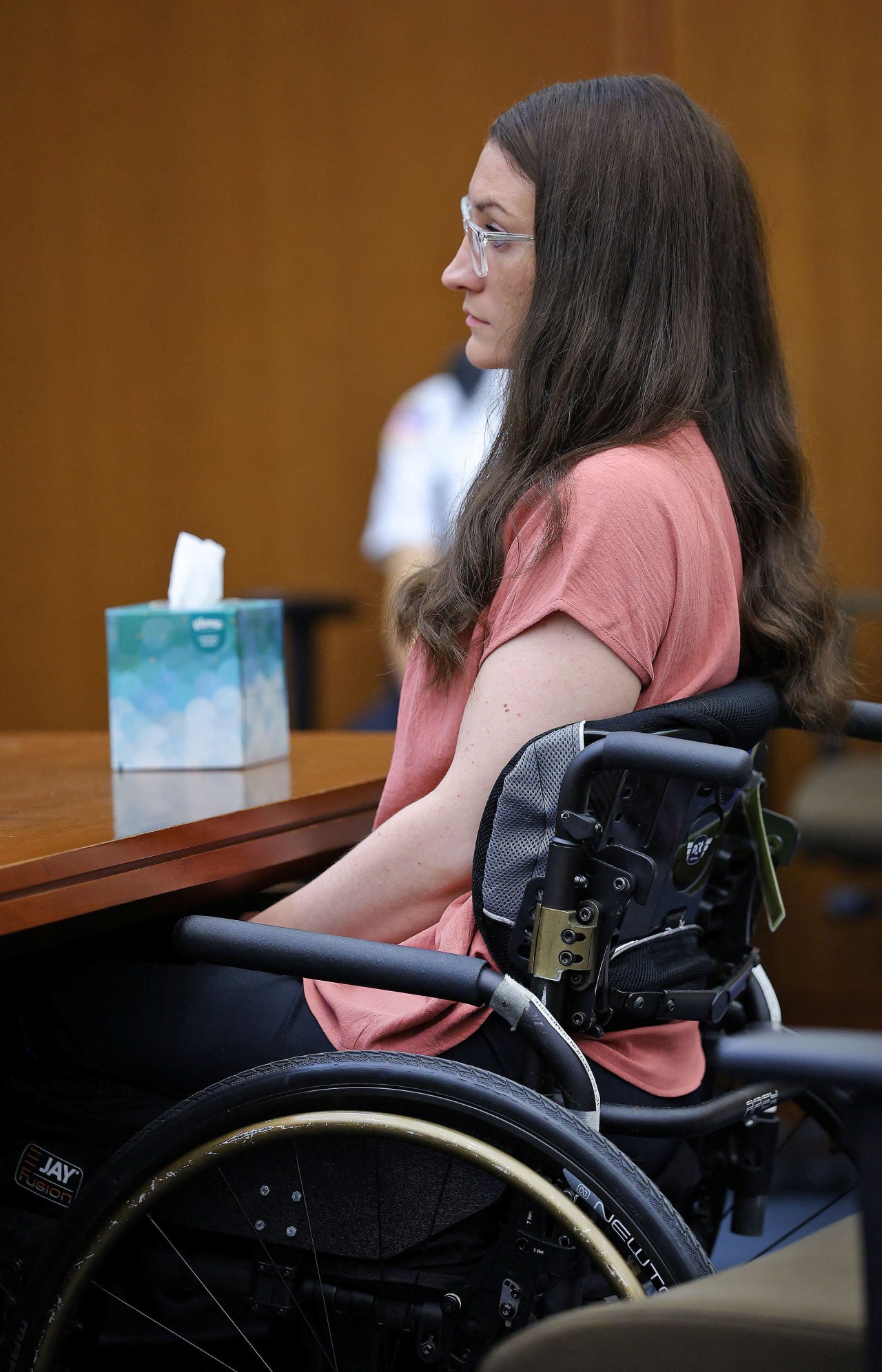 Woman with long brown hair and clear-framed glasses seated in a wheelchair at a courtroom desk, looking forward, viewed from the side.