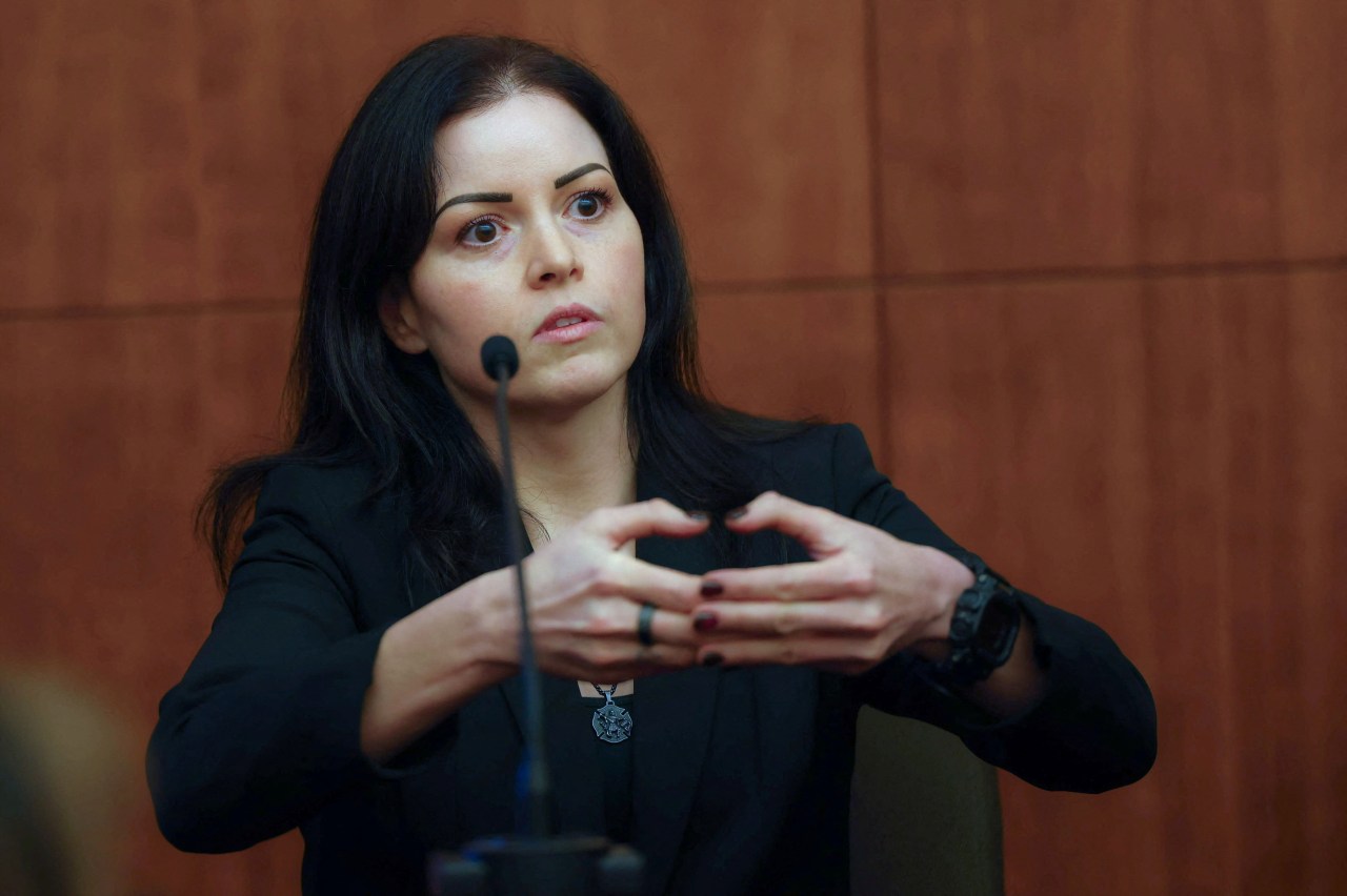 Dark-haired woman in a black blazer gesturing with both hands in front of a courtroom microphone while speaking.