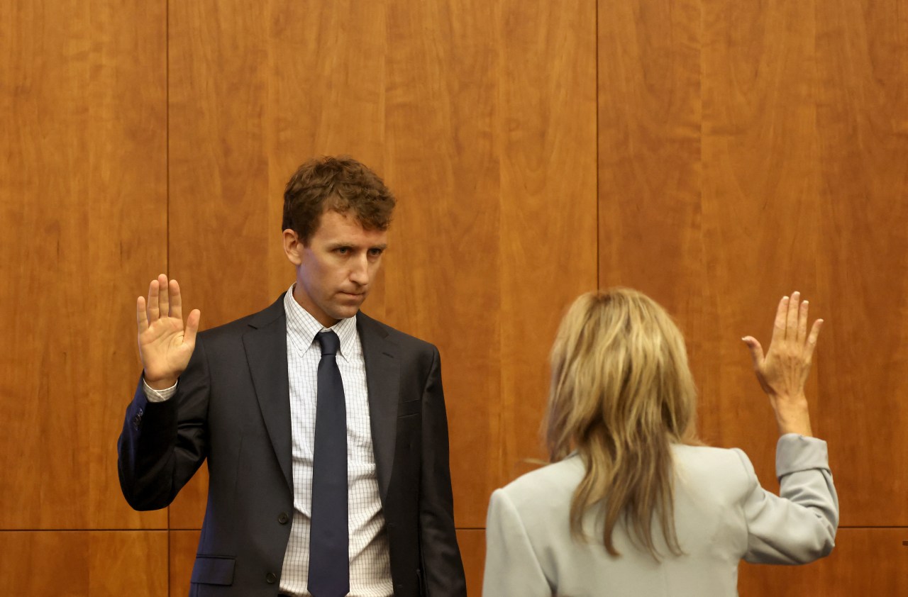 A man in a dark suit and blue tie raises his right hand to be sworn in, facing a woman with her back to the camera in a courtroom.