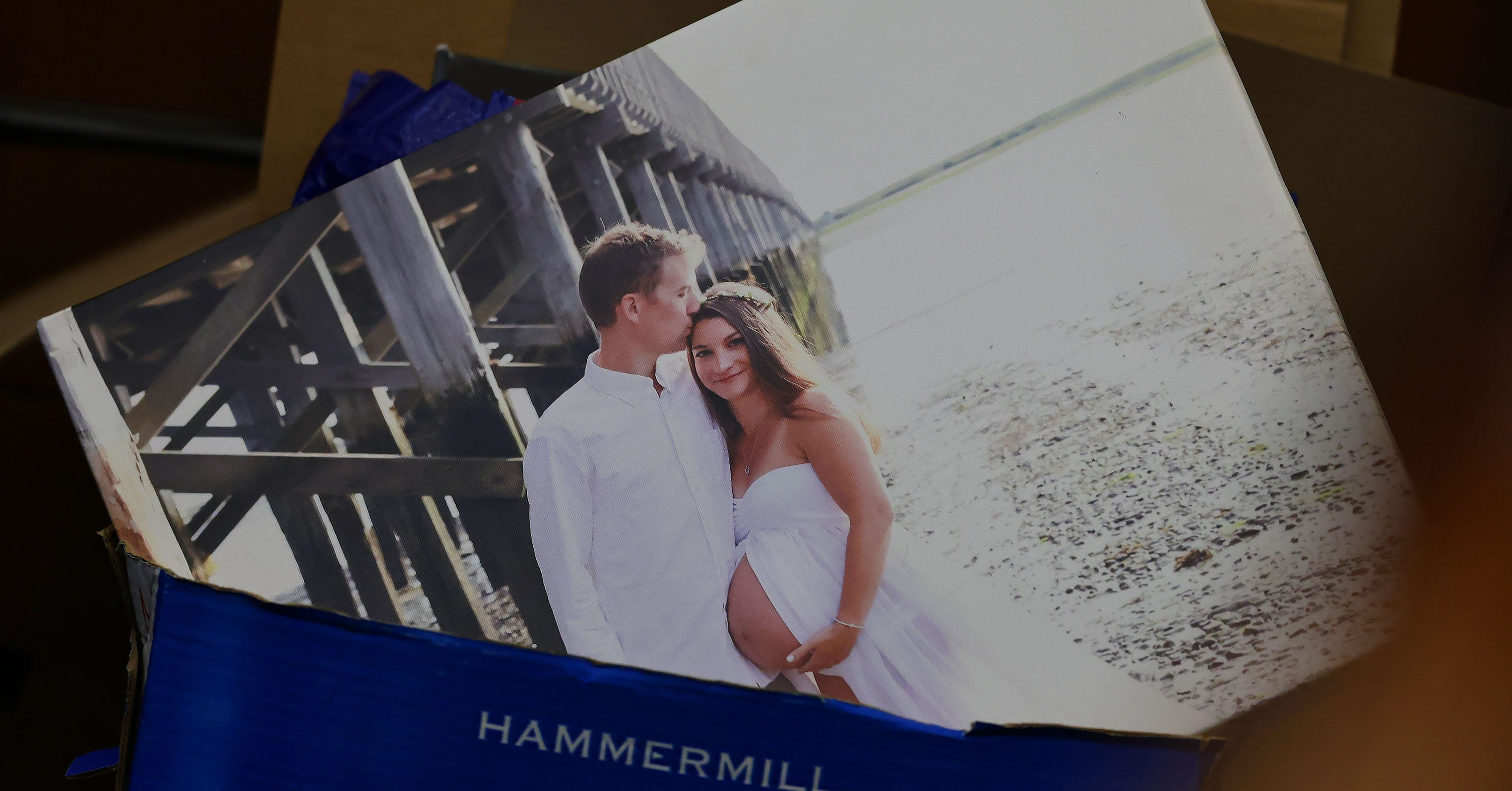 Printed maternity photo of a couple in white clothing near a wooden pier on a beach, resting on a Hammermill paper box.