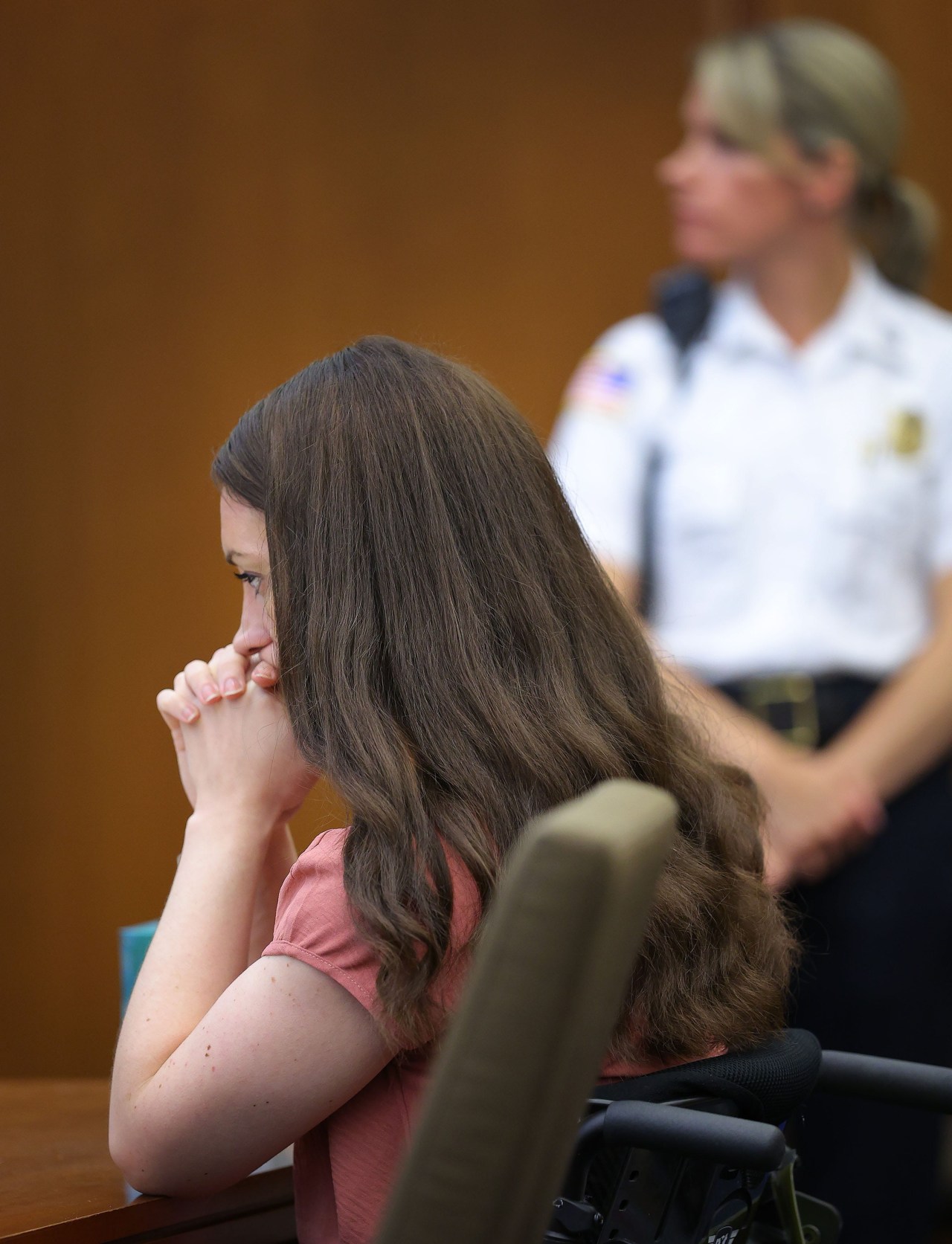 Woman with long brown hair in a pink top sits at a courtroom table with hands clasped over her mouth; a uniformed officer stands blurred in the background.