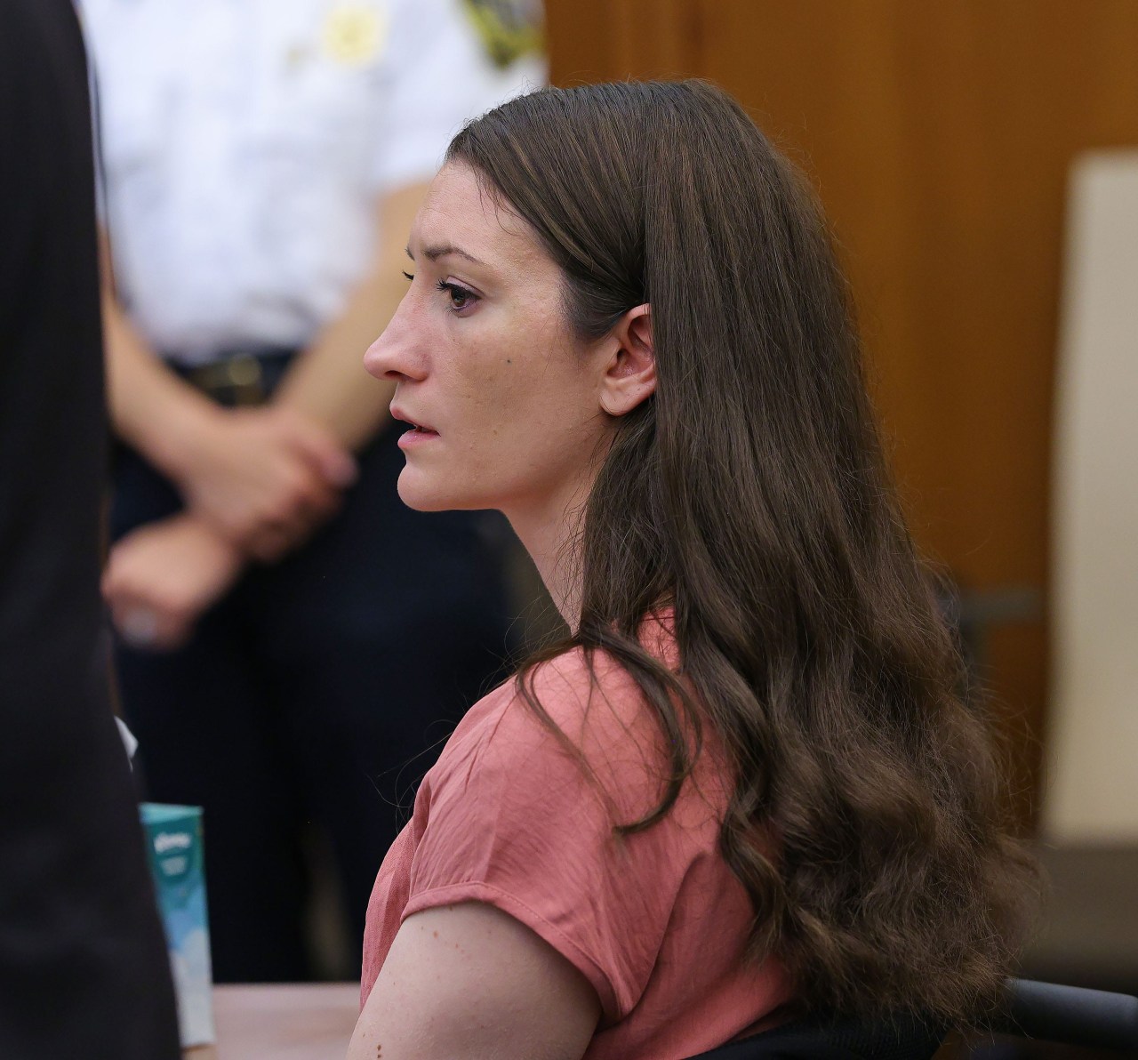 Side profile of a woman with long brown hair in a pink top seated in a courtroom, looking forward with a serious expression.