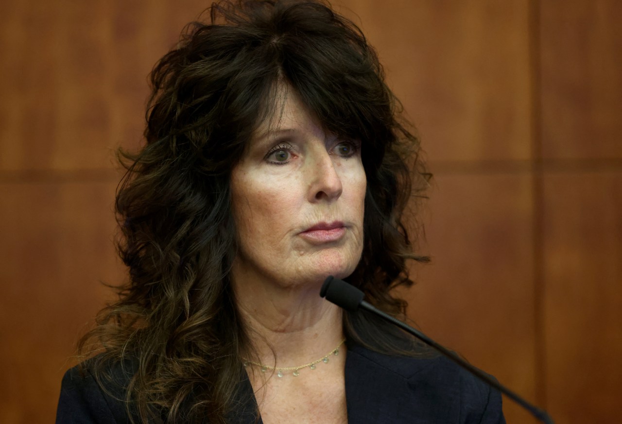 A middle-aged woman with dark curly hair in a navy blazer speaks at a microphone on a courtroom witness stand.