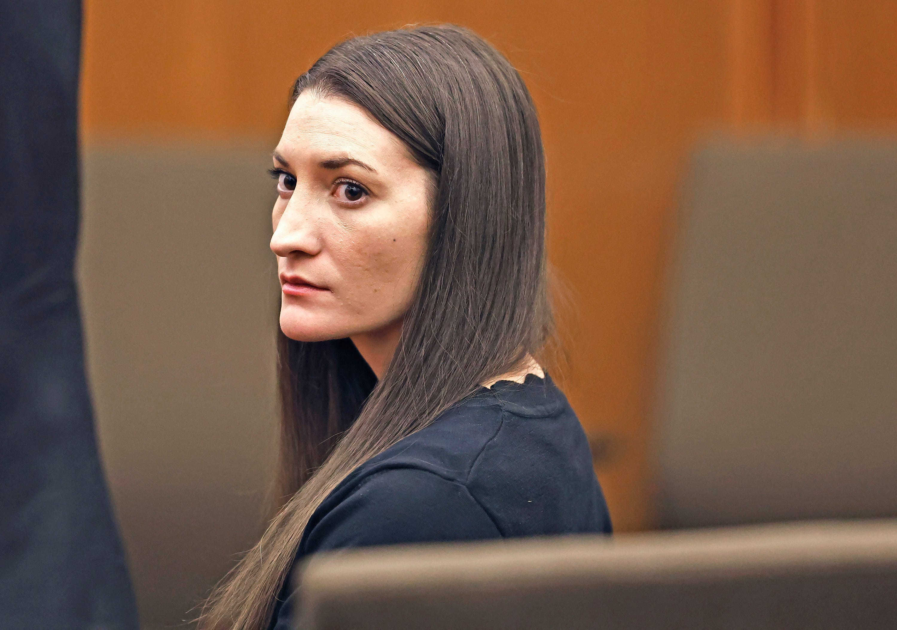 Woman with long dark hair in a black top seated in a courtroom, looking sideways toward the camera.