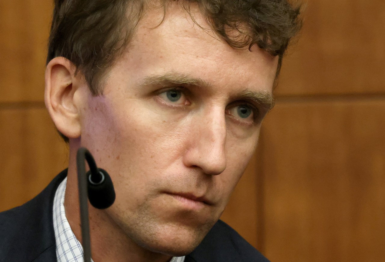 Close-up of a man with curly brown hair and blue eyes wearing a lapel microphone, seated in a courtroom witness stand.