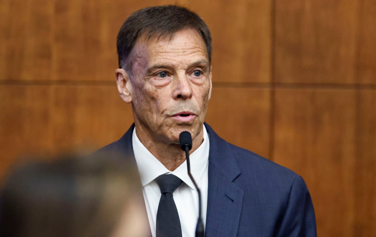 Middle-aged man in navy suit and black tie speaking into a microphone at a witness stand in a courtroom.