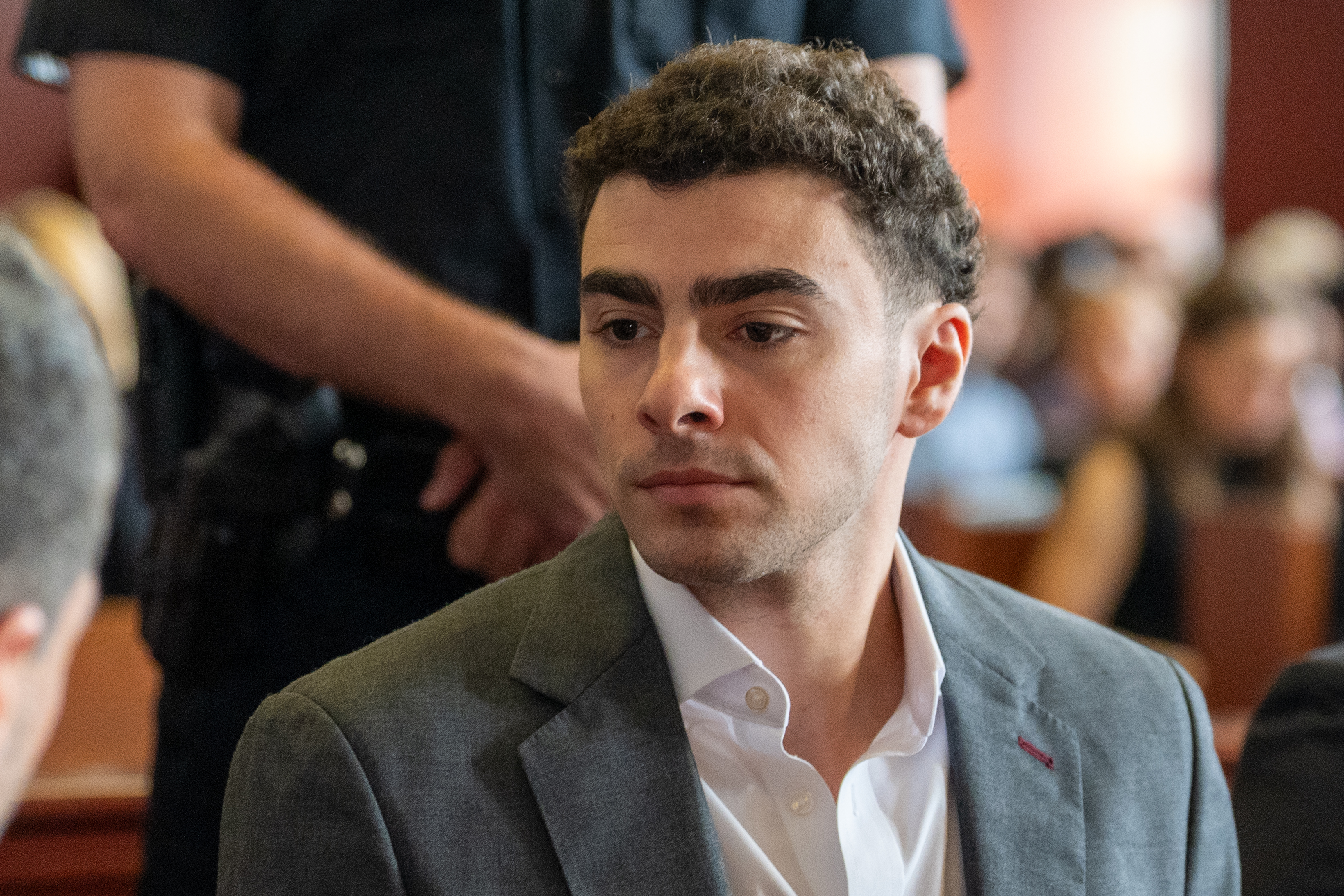 Young man in a gray blazer and white shirt seated in a courtroom, with a uniformed officer standing behind him.
