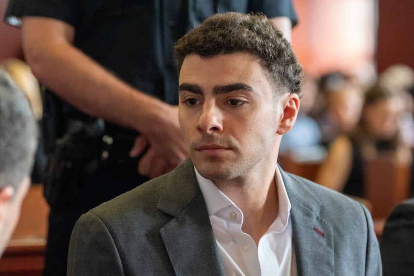 Young man in a gray blazer and white shirt seated in a courtroom, with a uniformed officer standing behind him.