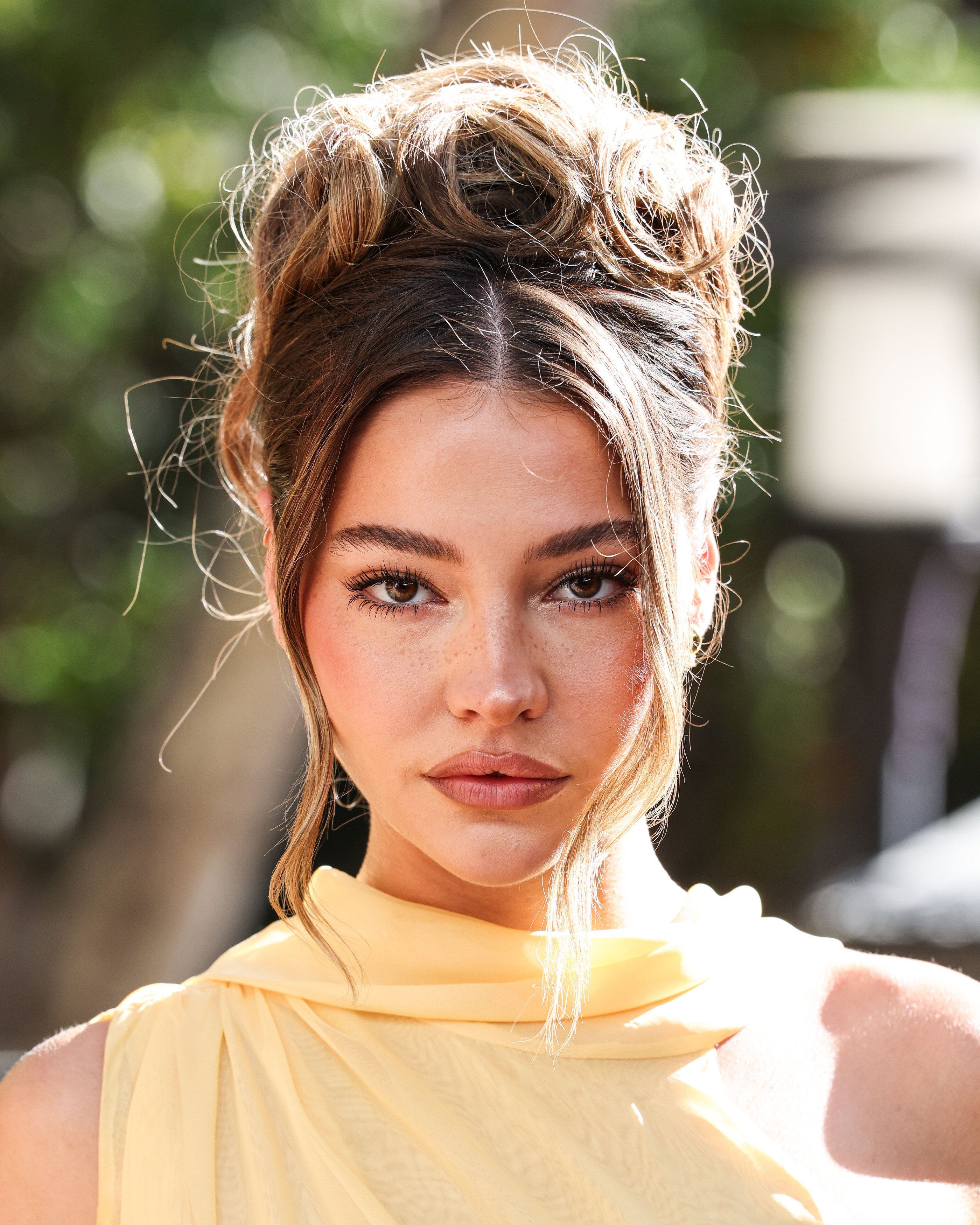 Close-up portrait of Madelyn Cline with a messy updo, wearing a pale yellow sleeveless top, outdoors at a fan event.