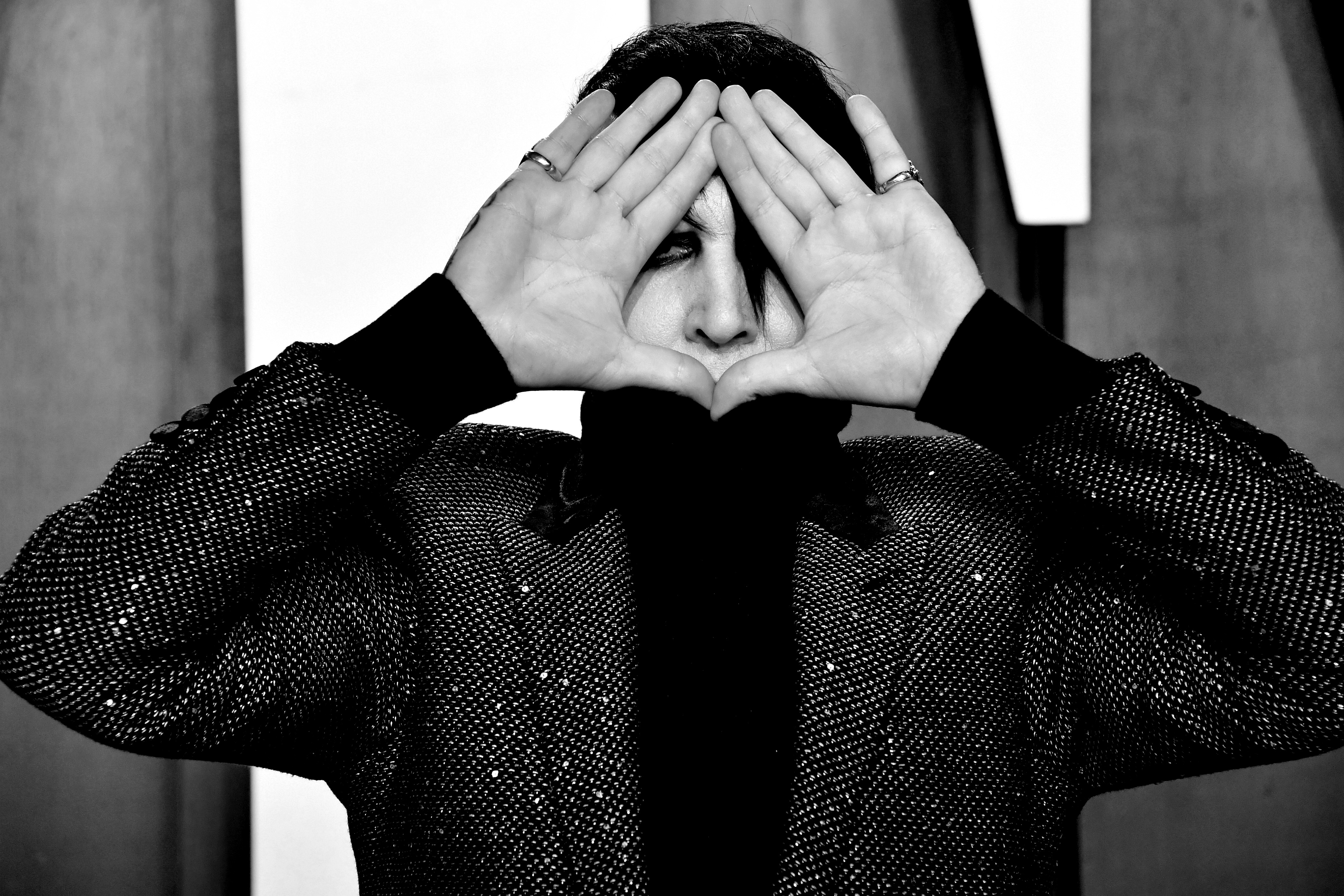 Black and white photo of Marilyn Manson holding both hands up to frame his face, wearing a sparkly dark jacket, at the 2020 Vanity Fair Oscar Party.