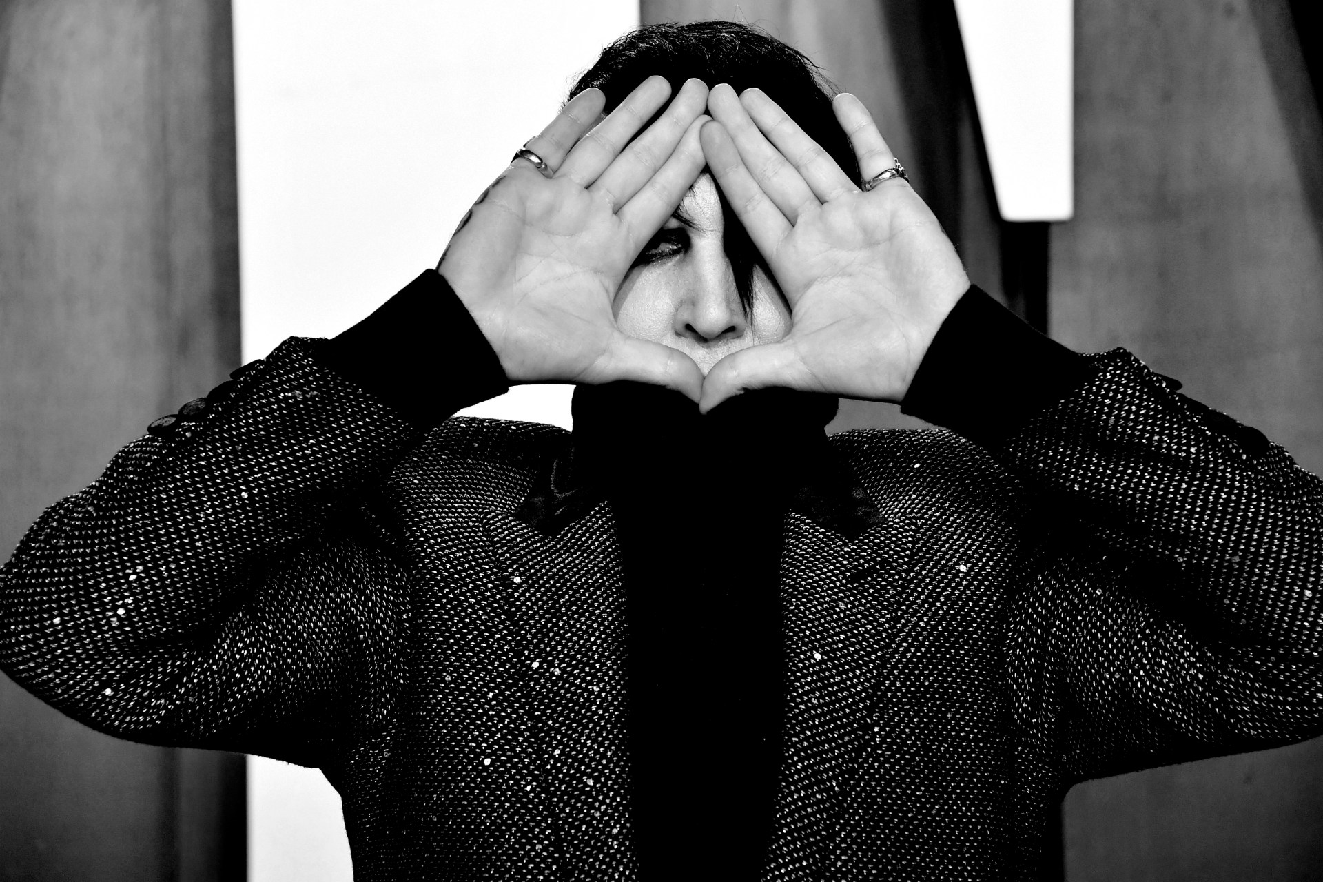Black and white photo of Marilyn Manson holding both hands up to frame his face, wearing a sparkly dark jacket, at the 2020 Vanity Fair Oscar Party.