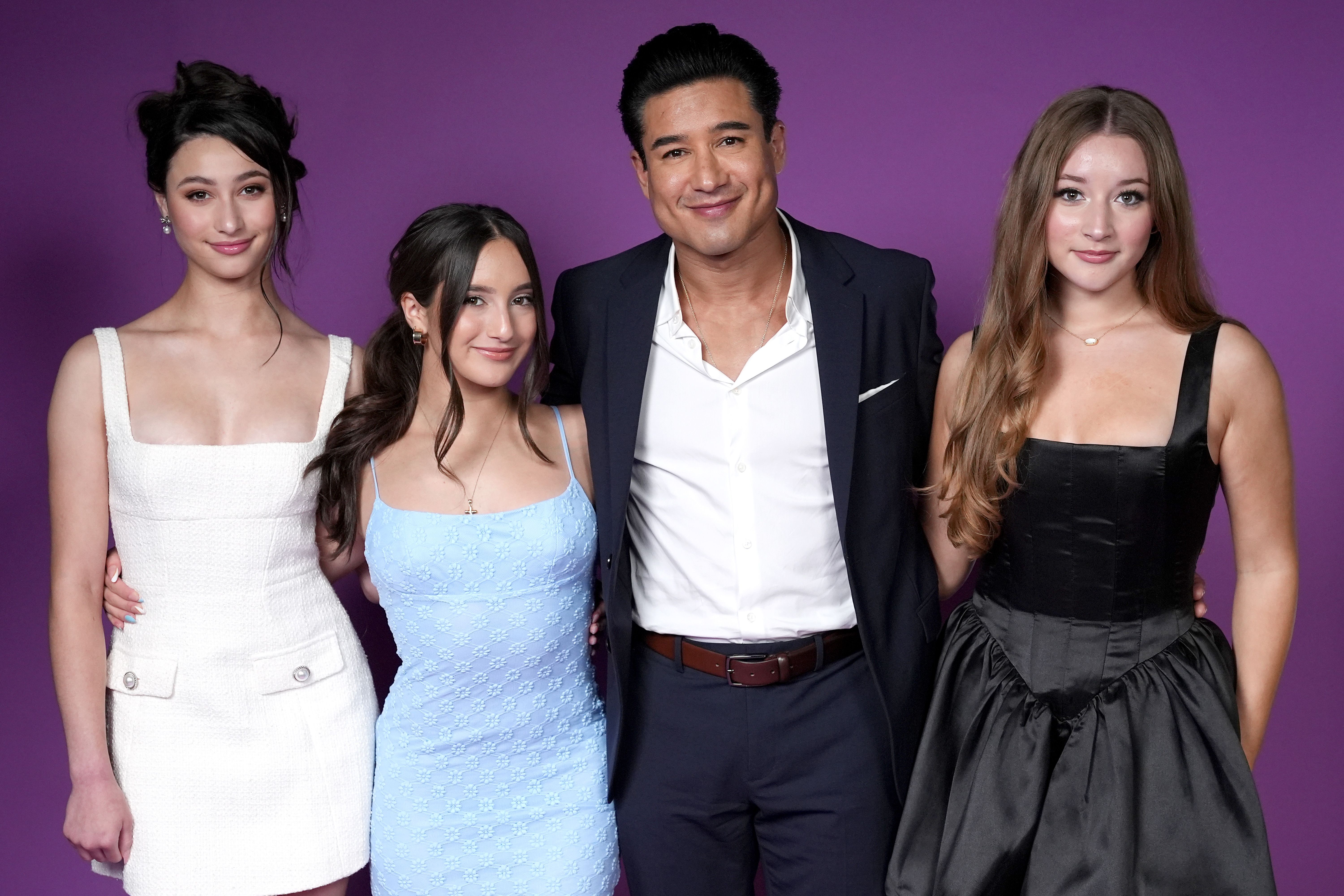 Mario Lopez in a navy suit stands with three young women — two in white and blue dresses on his left, one in a black dress on his right — against a purple background.