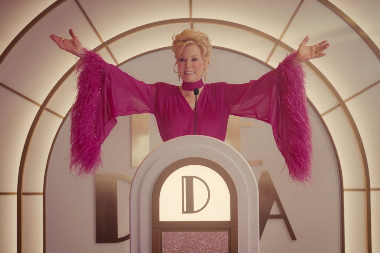 Woman in a hot-pink feathered gown standing at a decorative podium with arms outstretched, smiling broadly.