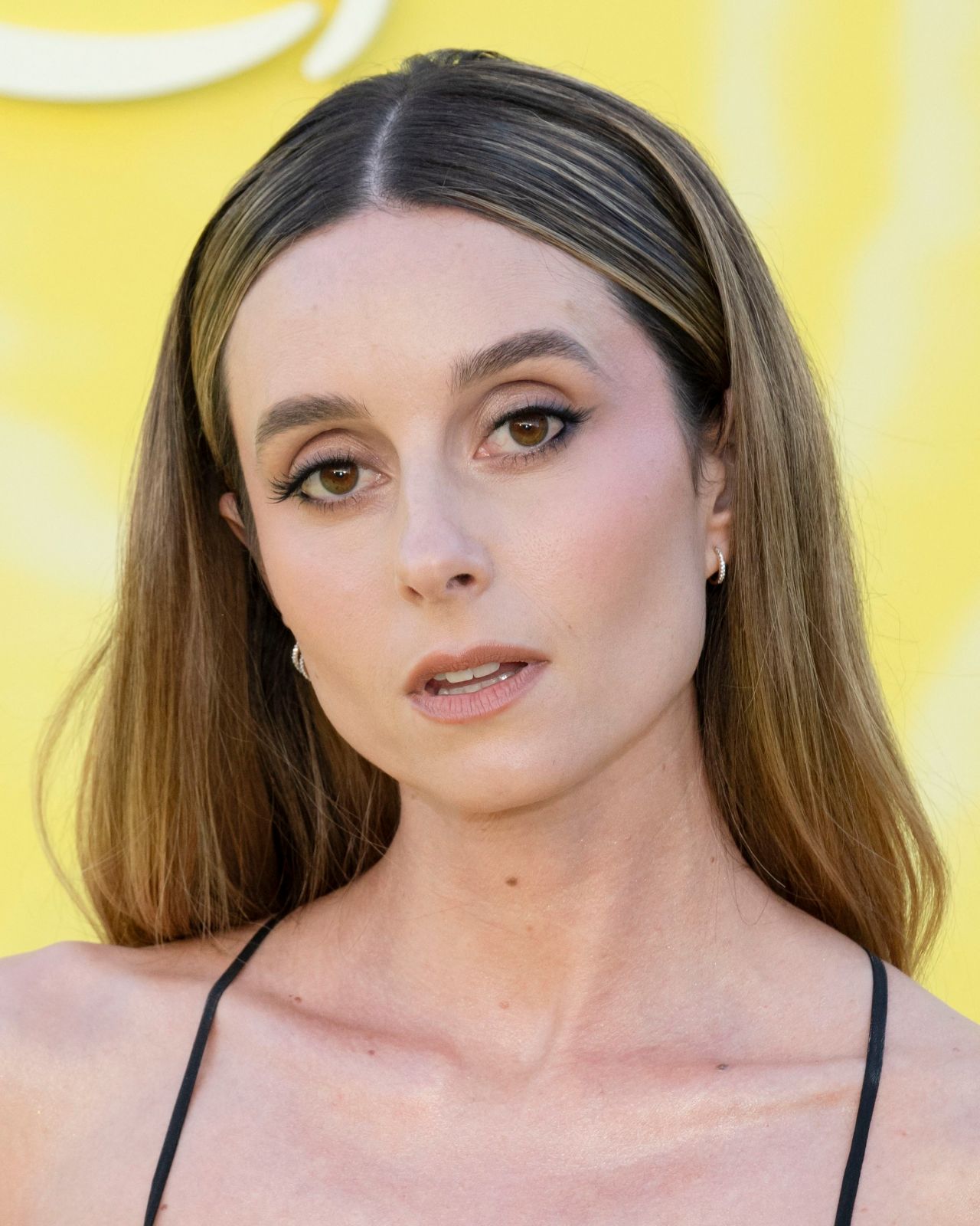 Wally Baram with long brown hair and hoop earrings poses against a yellow backdrop at a red carpet premiere.