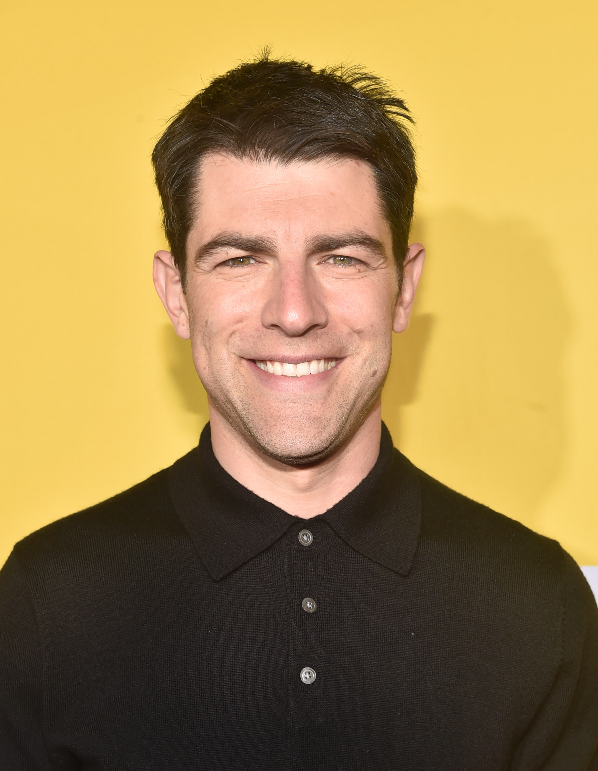 Max Greenfield smiling in a black polo shirt against a yellow backdrop at a movie premiere red carpet.