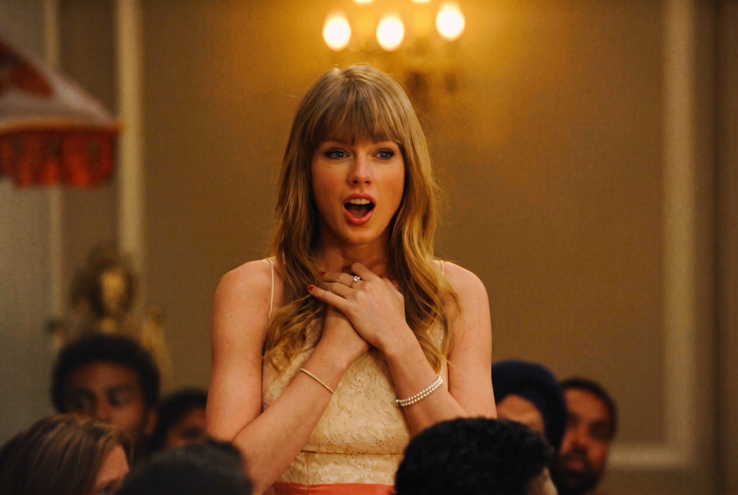 A blonde woman with bangs holds her hands to her chest with a surprised open-mouthed expression at an indoor party, surrounded by other guests.