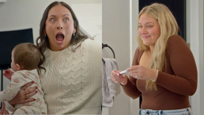 Woman in white cable-knit sweater holding a baby, mouth open in shock, reaching toward a pregnancy test on a bathroom counter.
