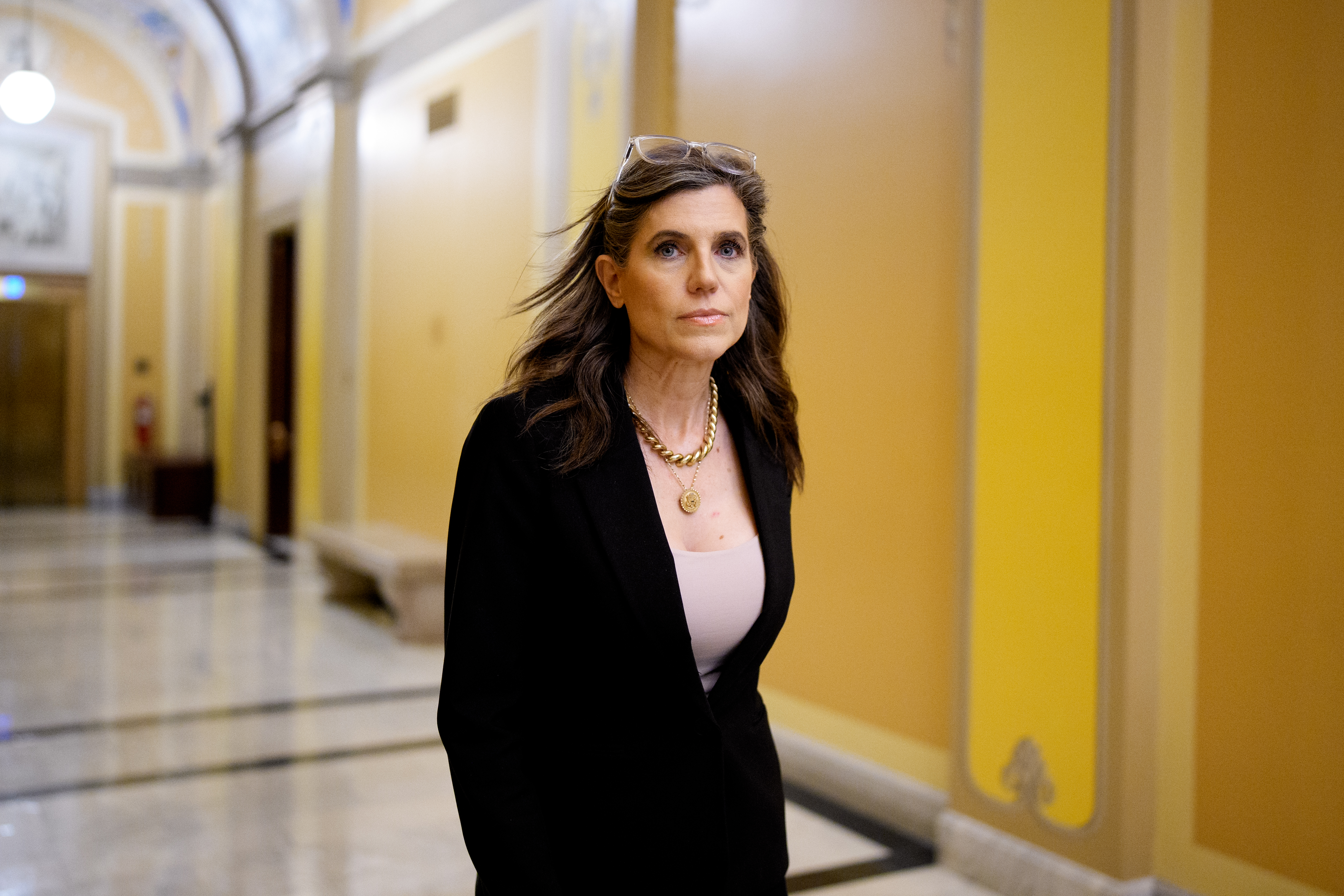 Rep. Nancy Mace in a black blazer and gold necklace walks through a yellow-walled Capitol hallway, glasses resting on her head.